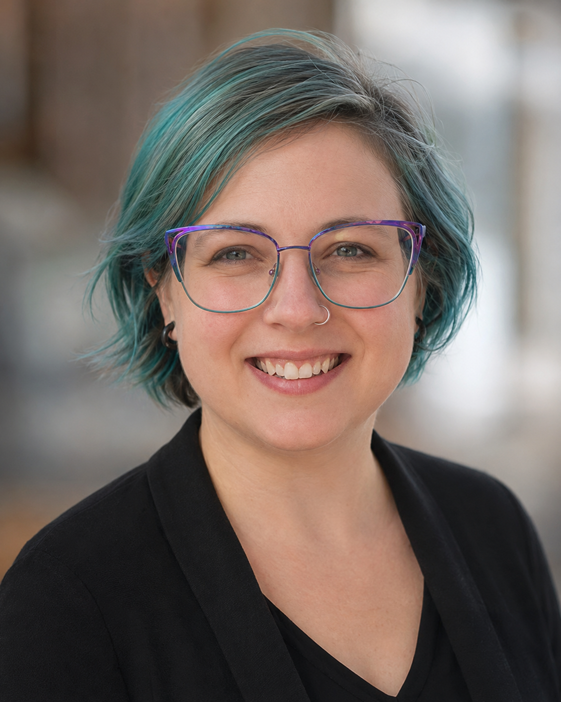 A woman with short, teal and gray hair, wearing large colorful glasses, smiling outdoors.