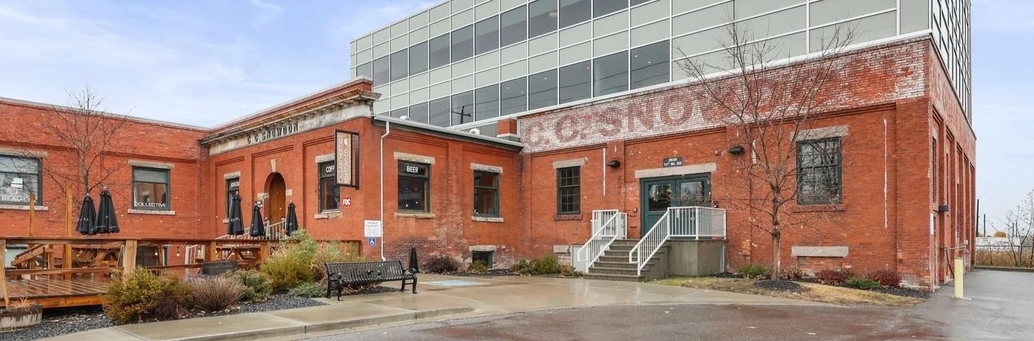 Red brick building with outdoor seating area, umbrellas, and signs advertising coffee, beer, and food. Small trees and a bench are in the front parking lot.