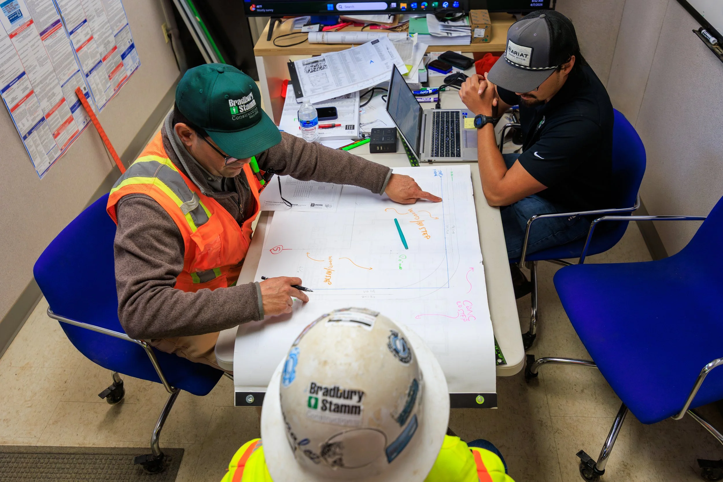People sitting at a cluttered desk discussing plans. One person wears a green cap and safety vest, pointing at papers, and a helmet in the foreground has a 'Bradbury Stam