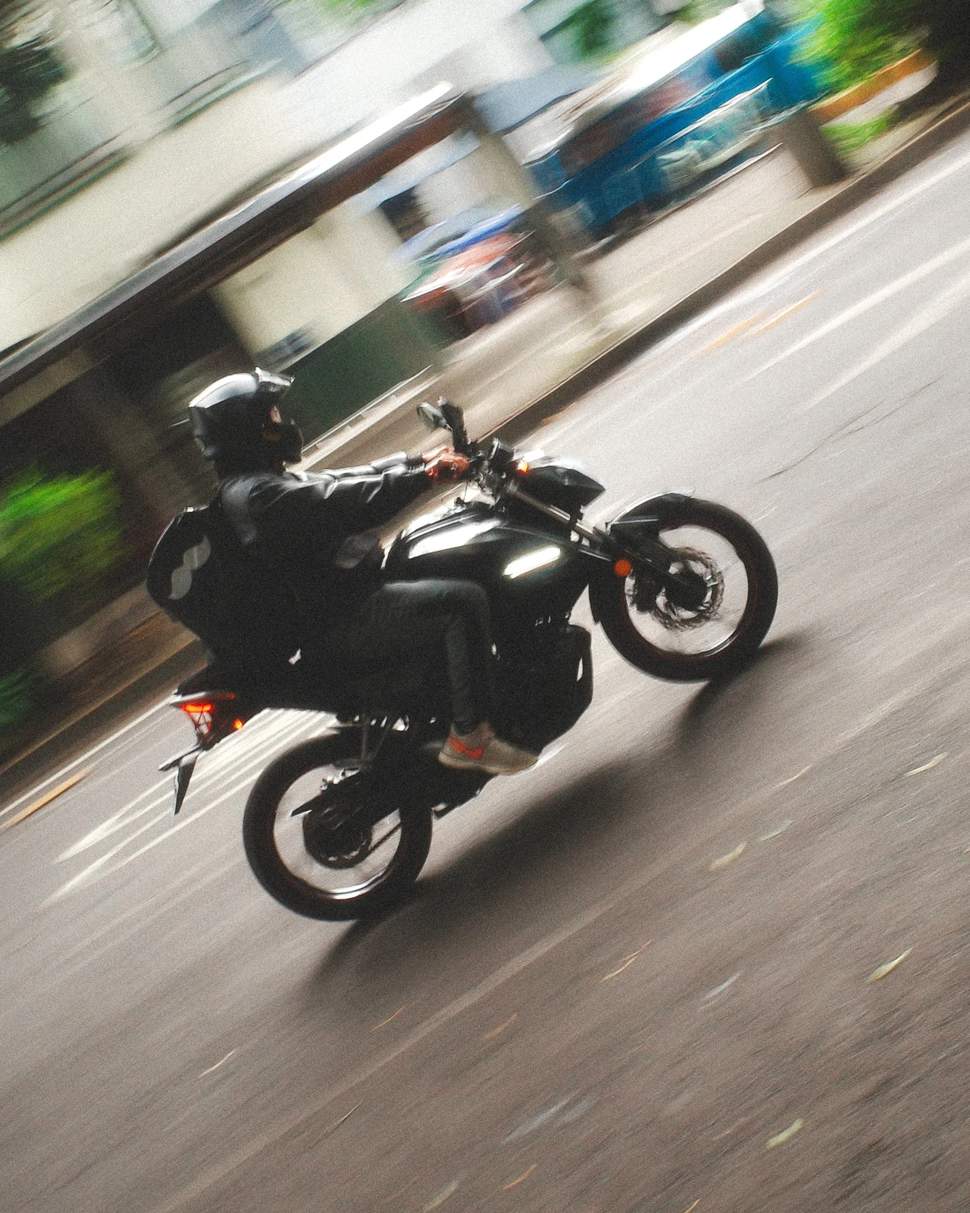 Blurred image of a person riding a motorcycle on a street, with the background showing a building and some greenery.