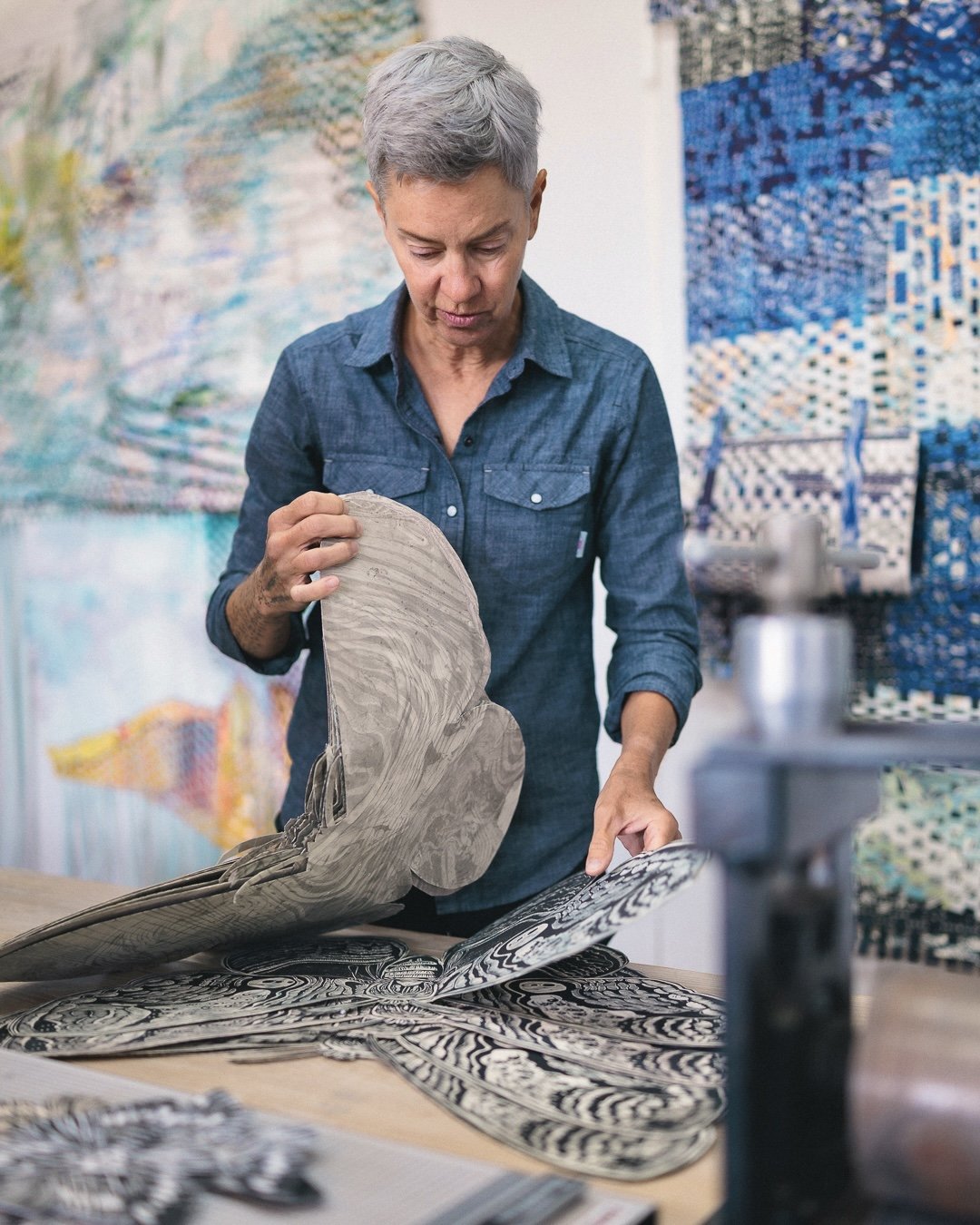 An artist examines a layered, abstract black-and-white artwork in a studio, surrounded by colorful paintings and art supplies.