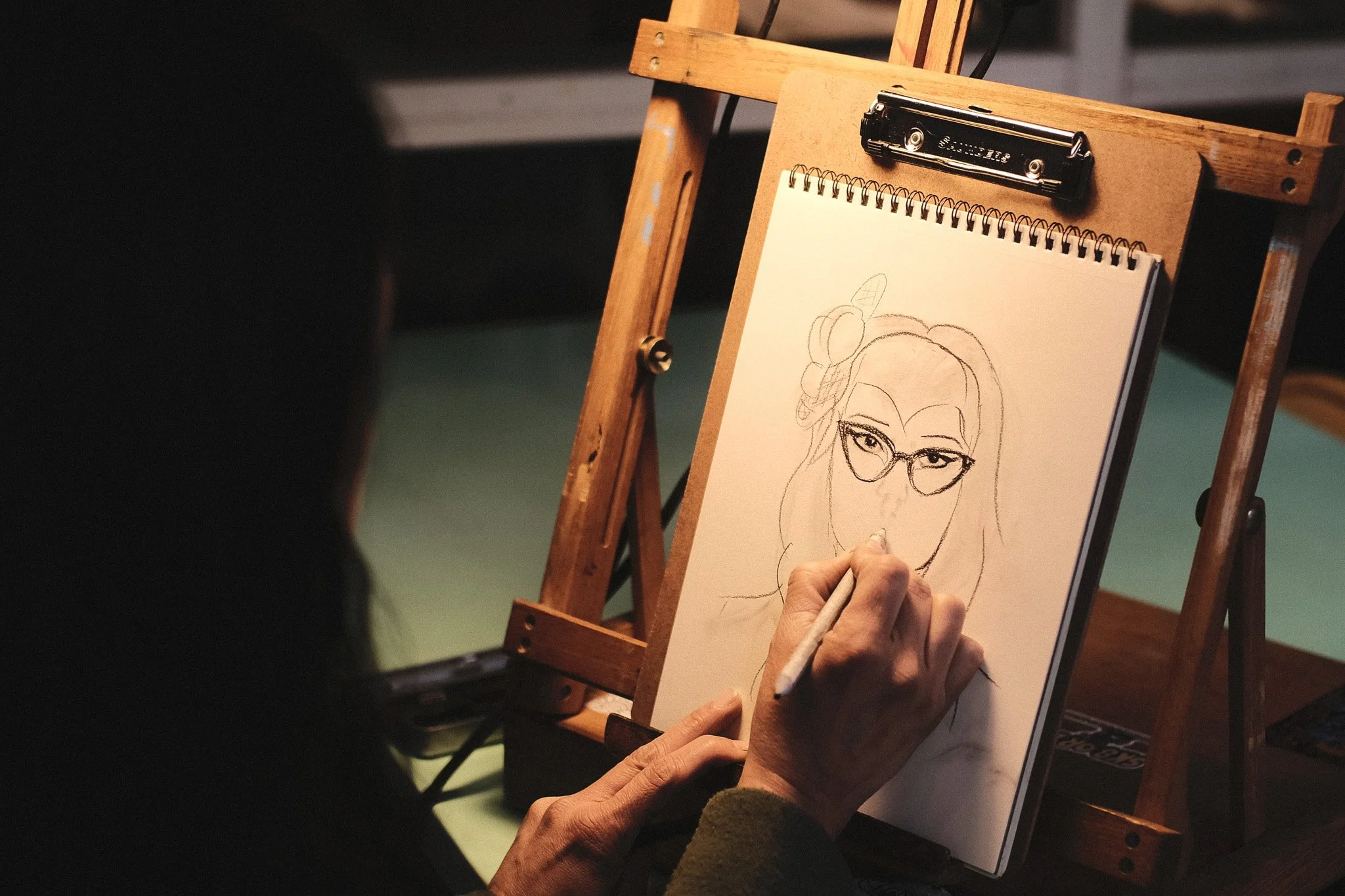 Person sketching a portrait of a woman with glasses on a notepad on an easel.