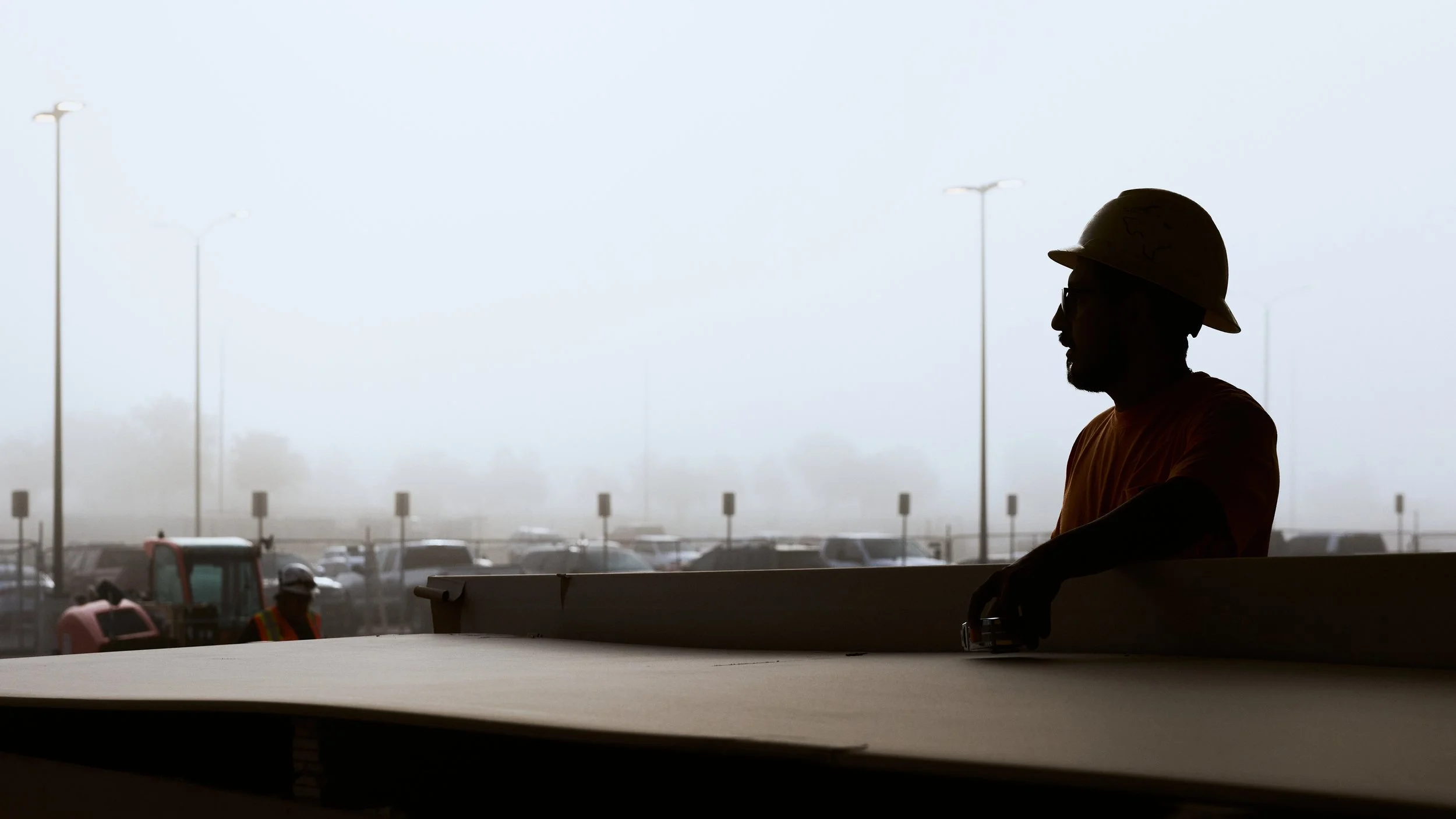 A construction worker wearing a helmet and orange shirt working at a counter with a foggy parking lot and cars in the background.