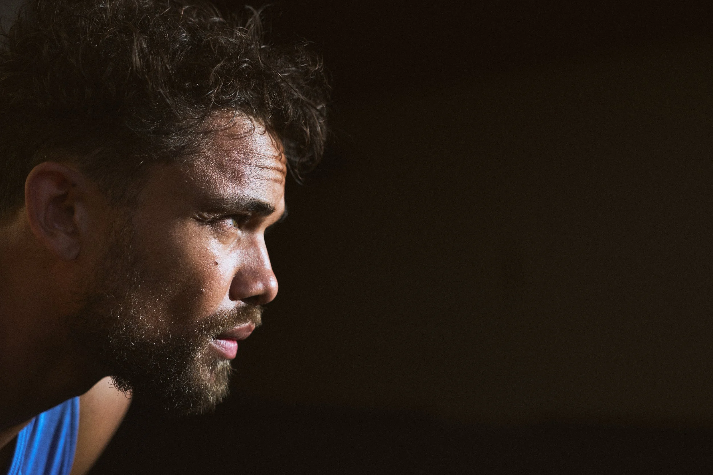 Close-up side profile of a man with curly dark hair, a beard, and wearing a blue shirt, looking to the left against a black background.