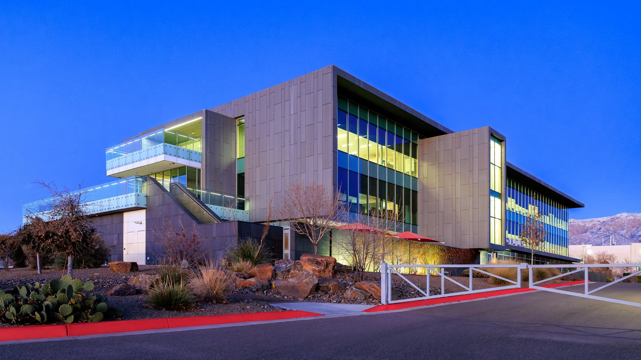 Modern office building with glass windows and concrete exterior, illuminated at dusk, with desert landscaping in the foreground and mountains in the background.
