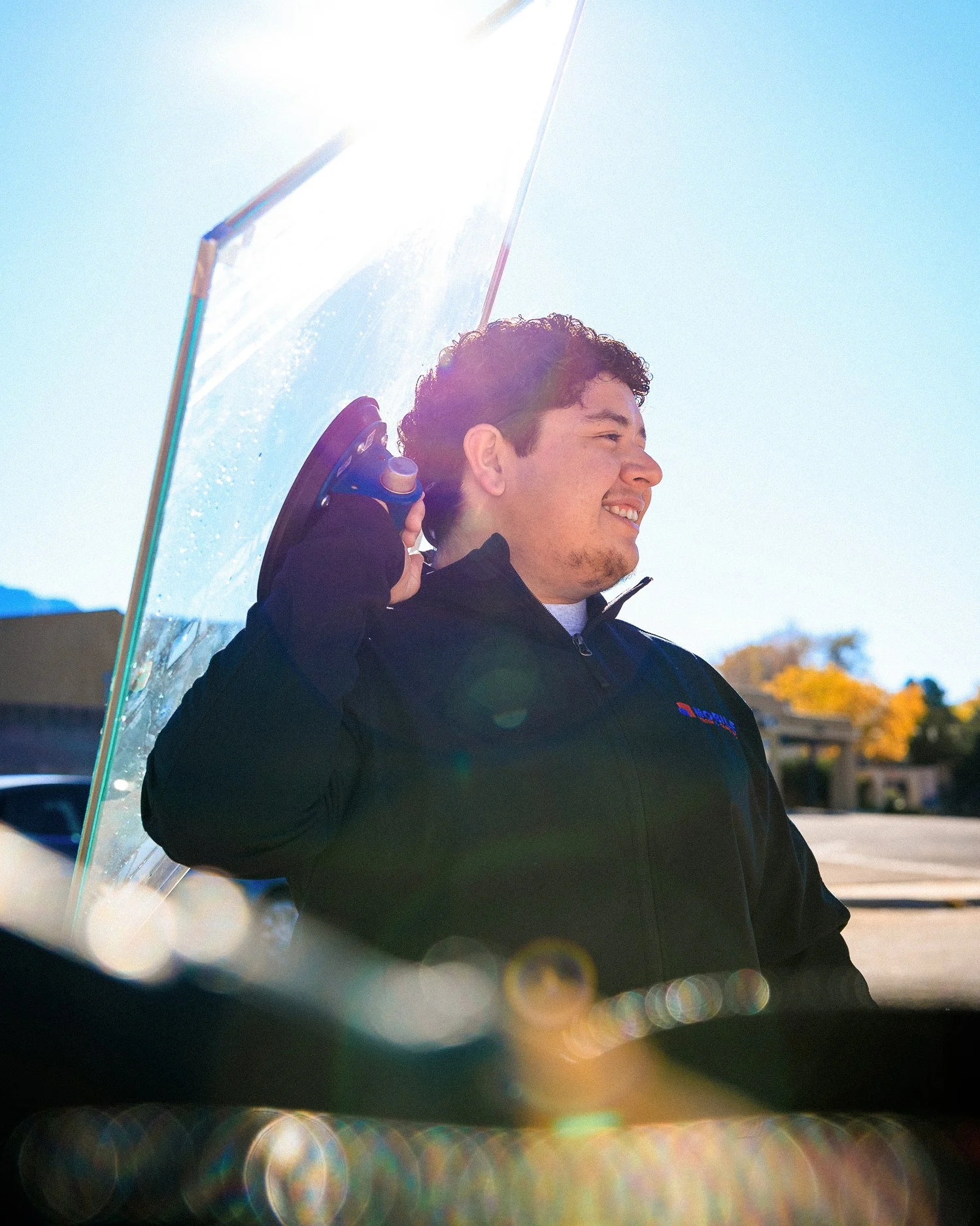 A young man with curly hair smiling and holding a large piece of glass or plastic behind his head, with sunlight behind him and a clear blue sky.
