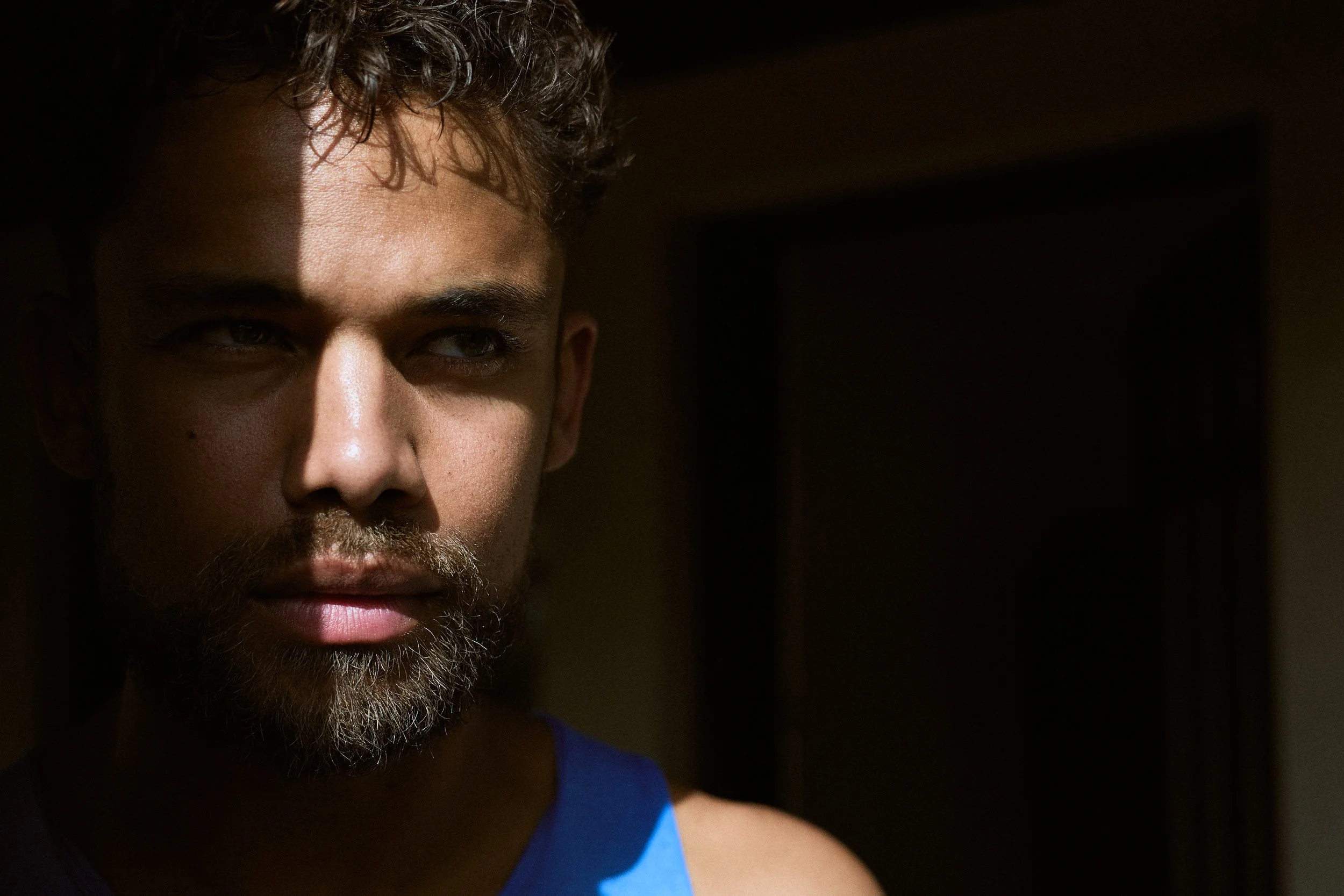 A close-up of a man with curly hair and a beard, looking into the distance in a dimly lit room.