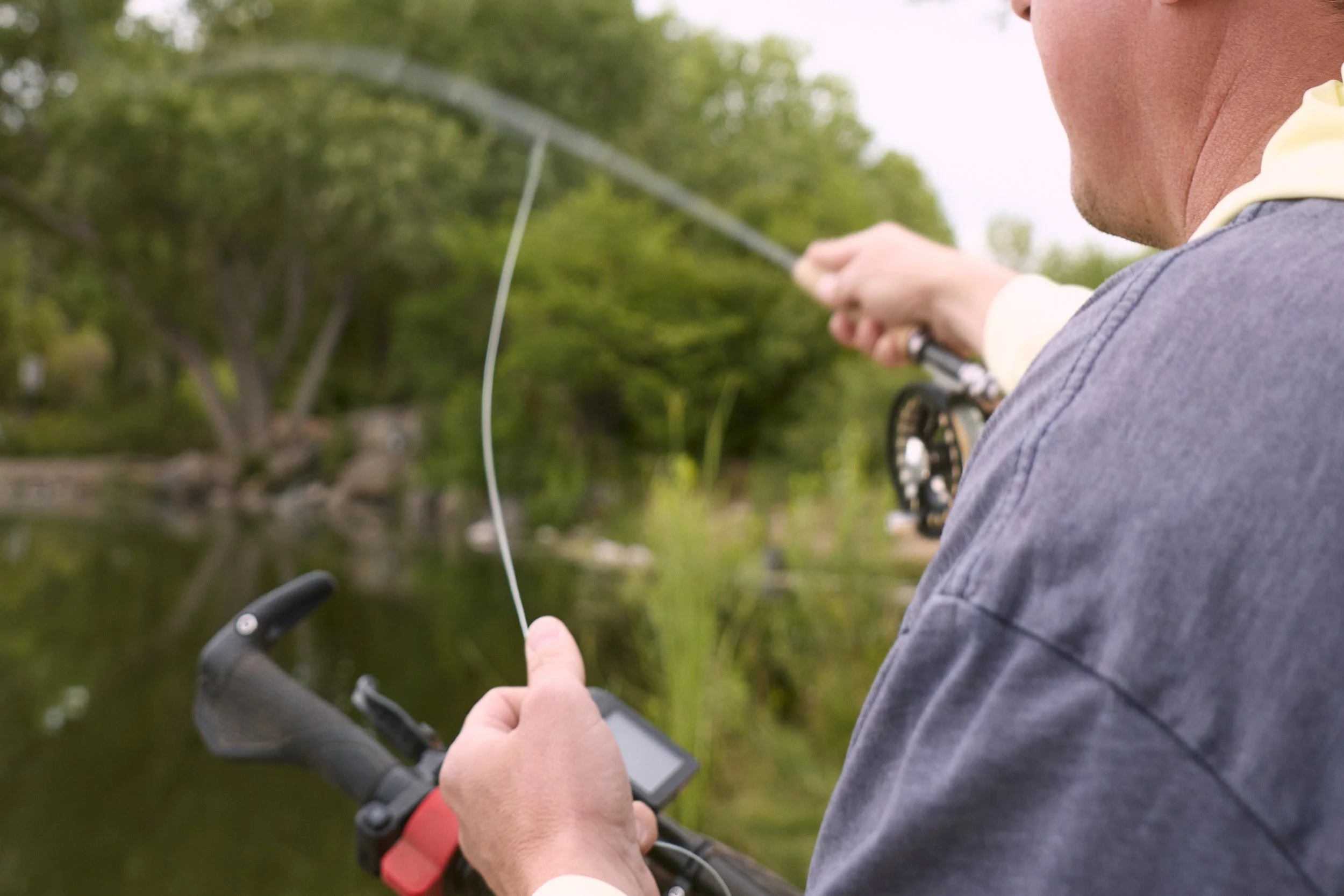 Close-up of a person’s hand holding a remote control device, pointing at another person fishing in a boat on a green, tree-lined body of water.