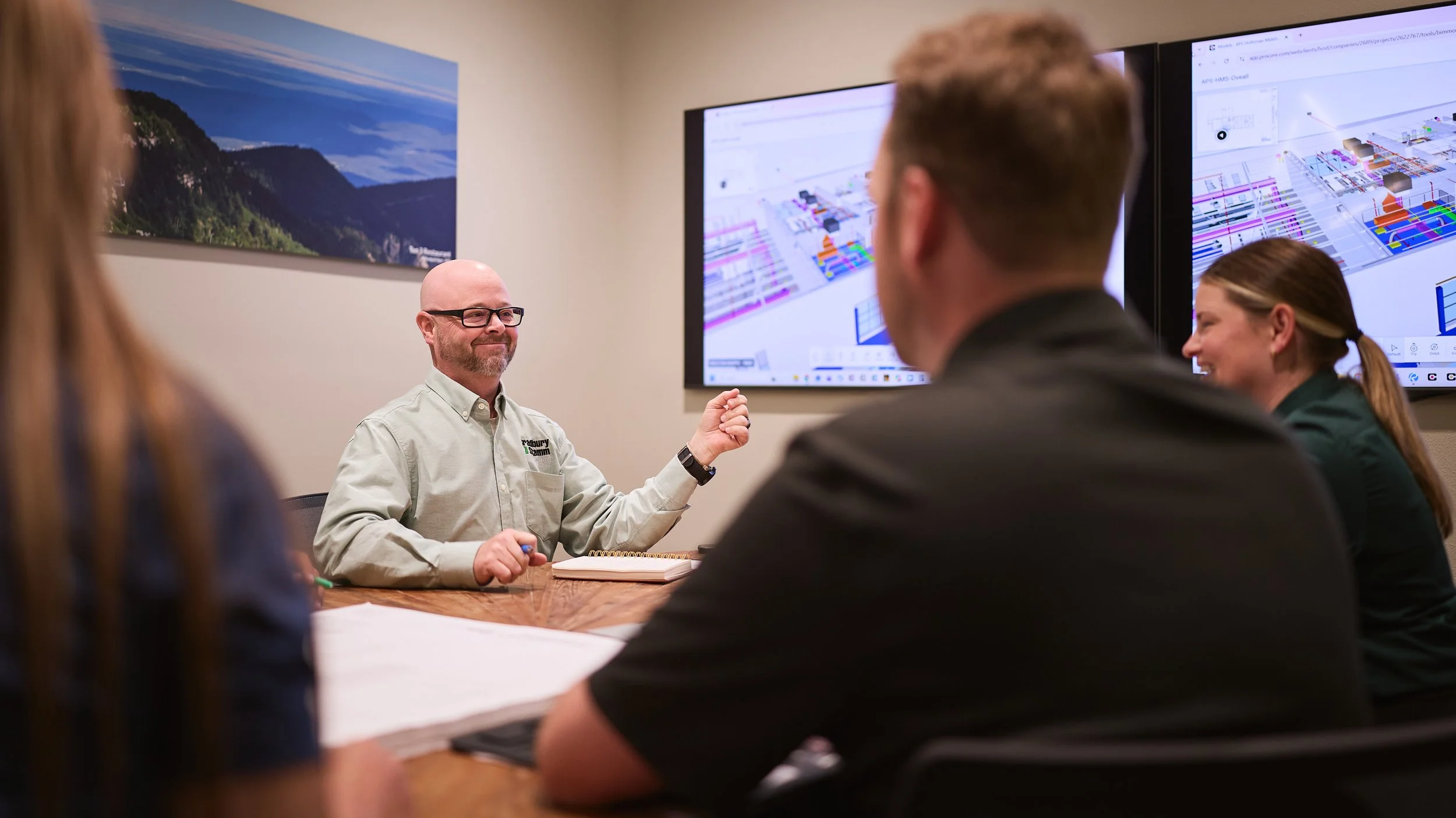 A group of people in a meeting room, watching a presentation on two large screens showing detailed diagrams or plans, with a man in a light-colored shirt speaking and gesturing.