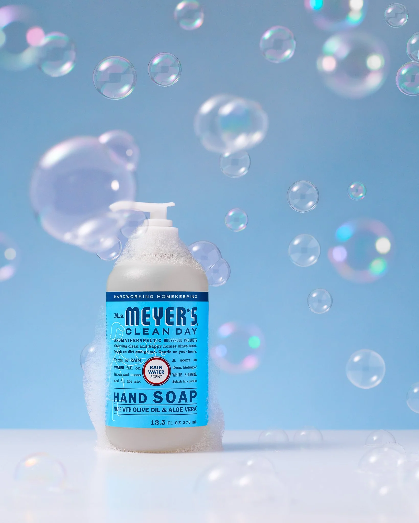 A bottle of Mrs. Meyer's Clean Day hand soap with a label indicating it is aromatic, made with olive oil and aloe vera, and contains 12.5 fluid ounces. The background features soap bubbles and a light blue surface.