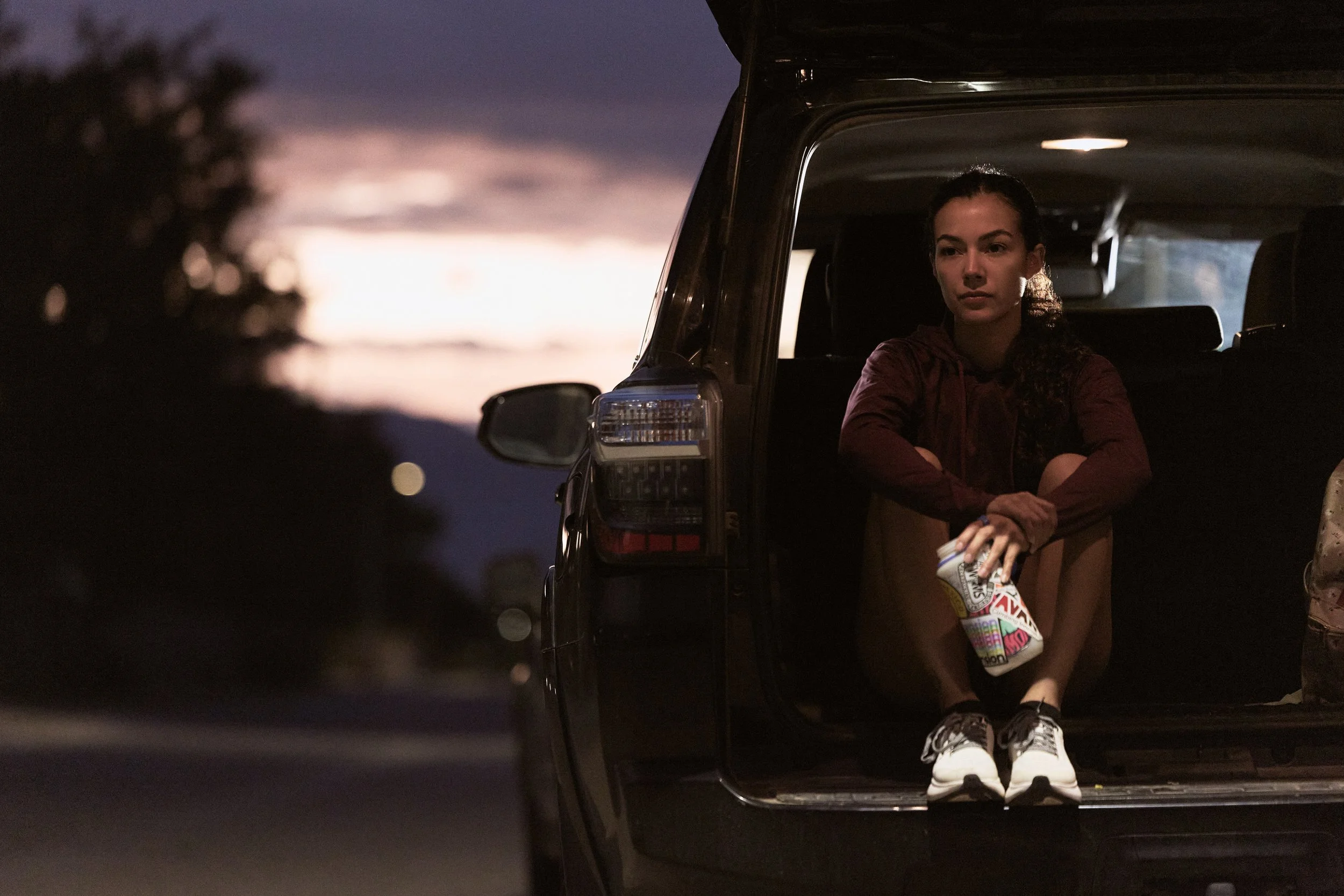 A young woman sitting in the open back of a vehicle at dusk, holding a snack bag, with mountains and a colorful sky in the background.