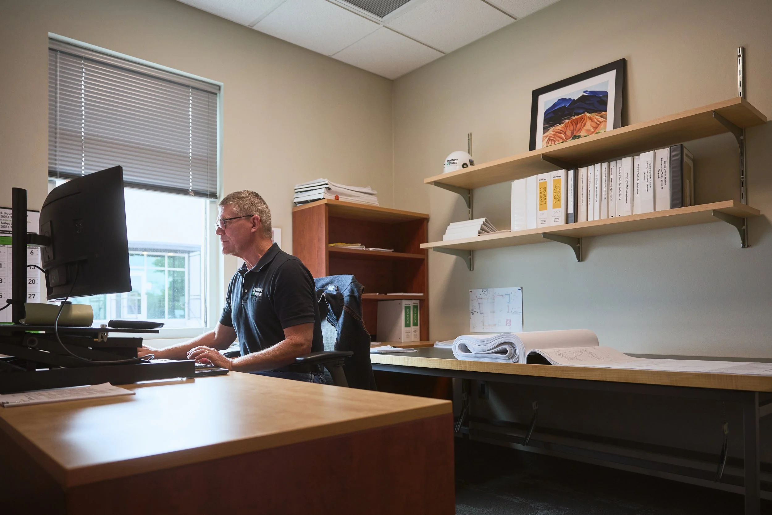 A man working at a desk in an office, with a computer monitor, paperwork, and a window with blinds behind him.