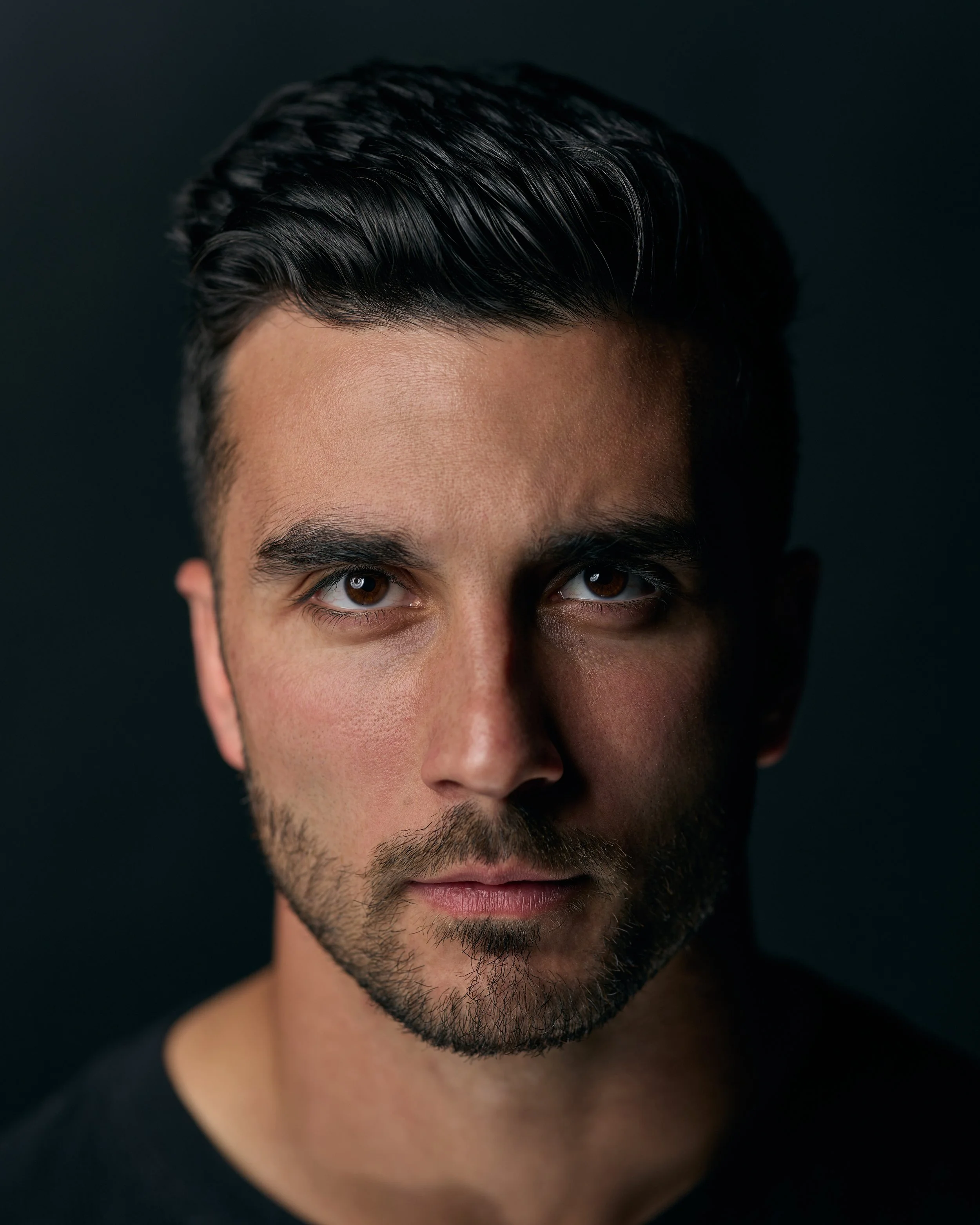 Close-up of a man's face, showing part of his eyes, nose, and mouth, with dark hair styled back and a beard, against a dark background.
