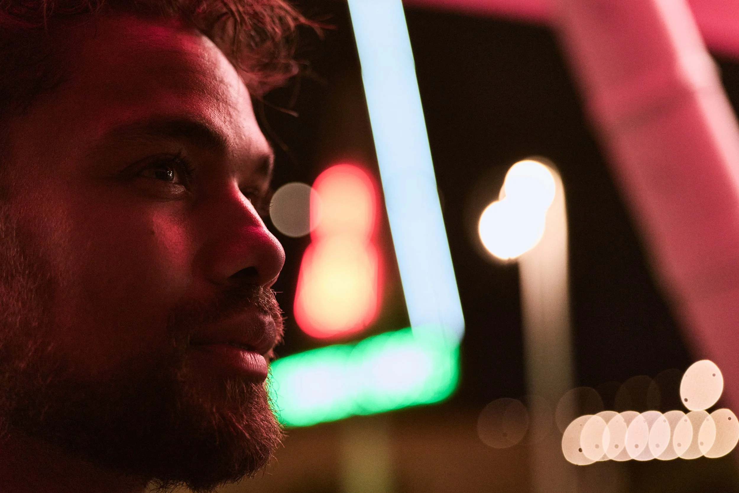Close-up of a man's face at night, illuminated by colorful neon lights and bokeh effects.
