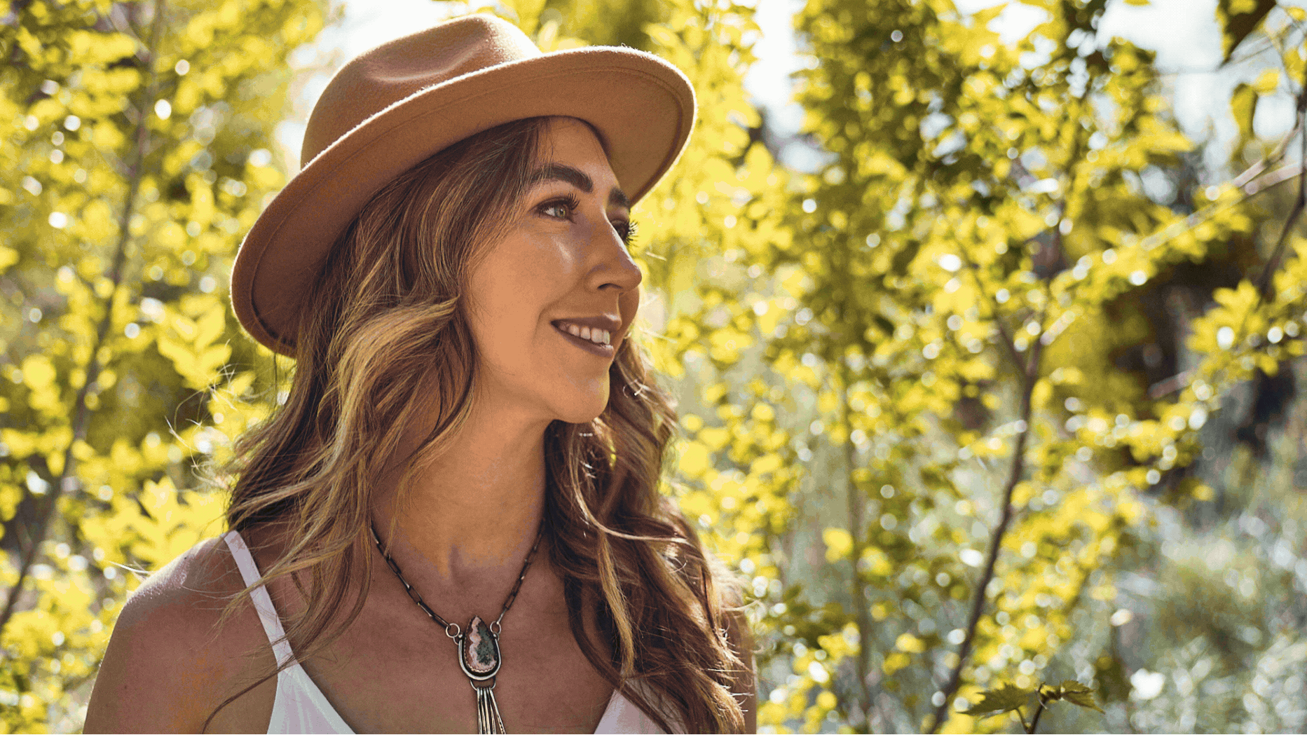 A woman with long wavy brown hair wearing a tan hat, a white tank top, and a necklace with a large stone pendant, smiling outdoors with yellow-green autumn leaves in the background.