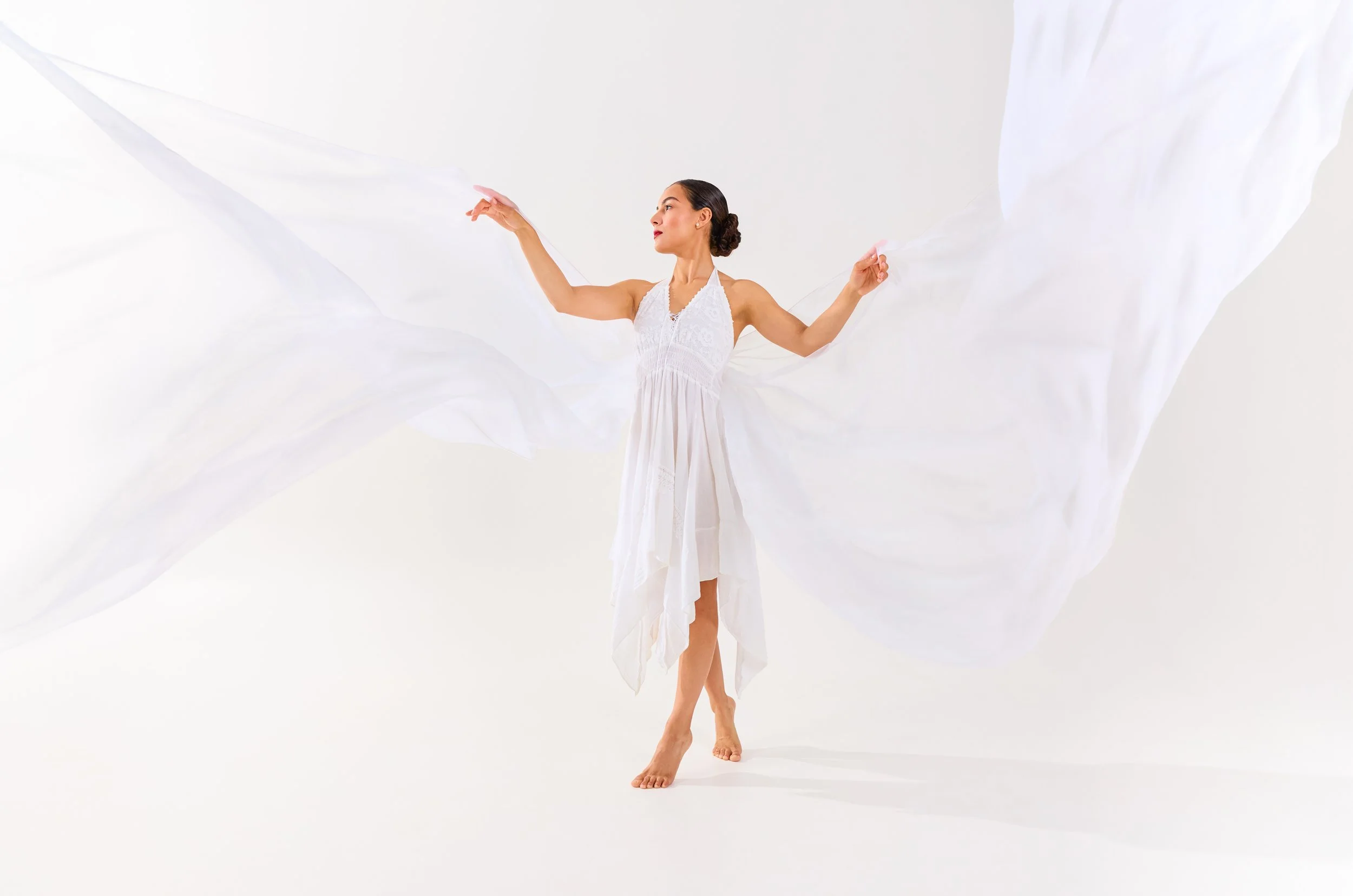 A woman dressed in a white gown with flowing fabric, standing on one foot with her arms outstretched, against a plain white background.
