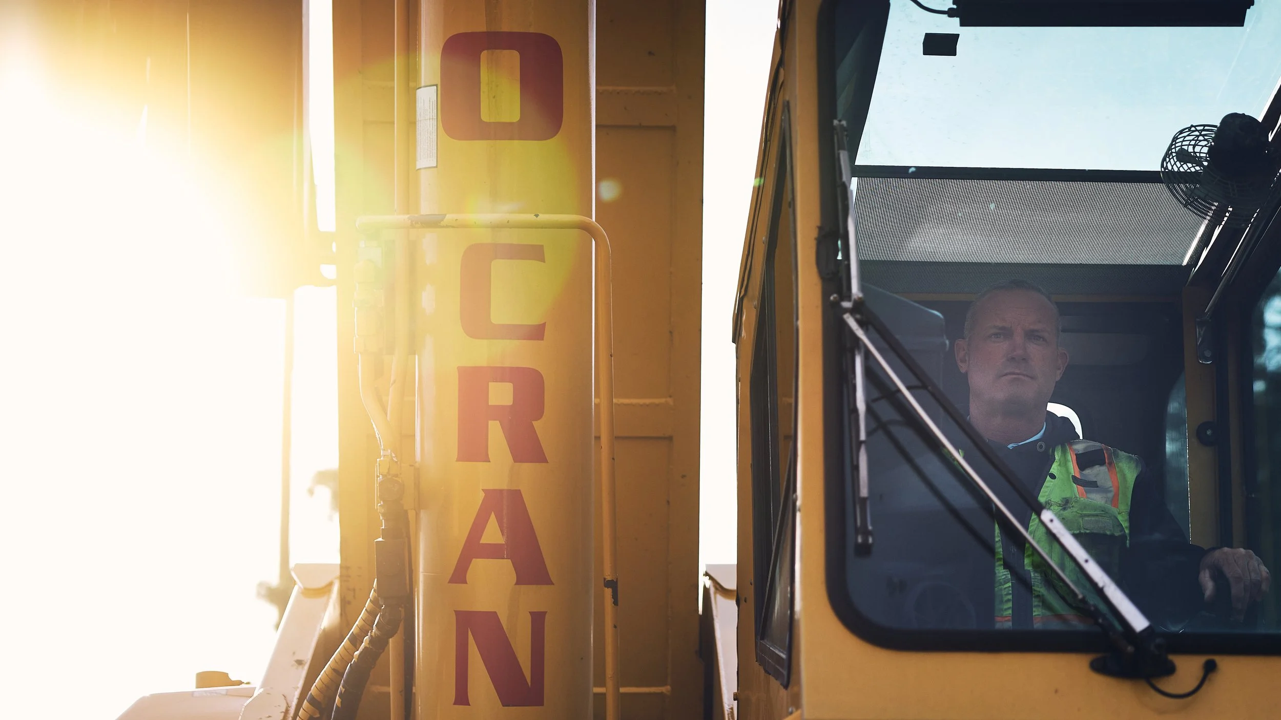 A person driving a large construction vehicle, possibly an excavator or loader, visible through the vehicle's window. The vehicle has a yellow exterior with a vertical caution sign, and bright sunlight reflects off the equipment.