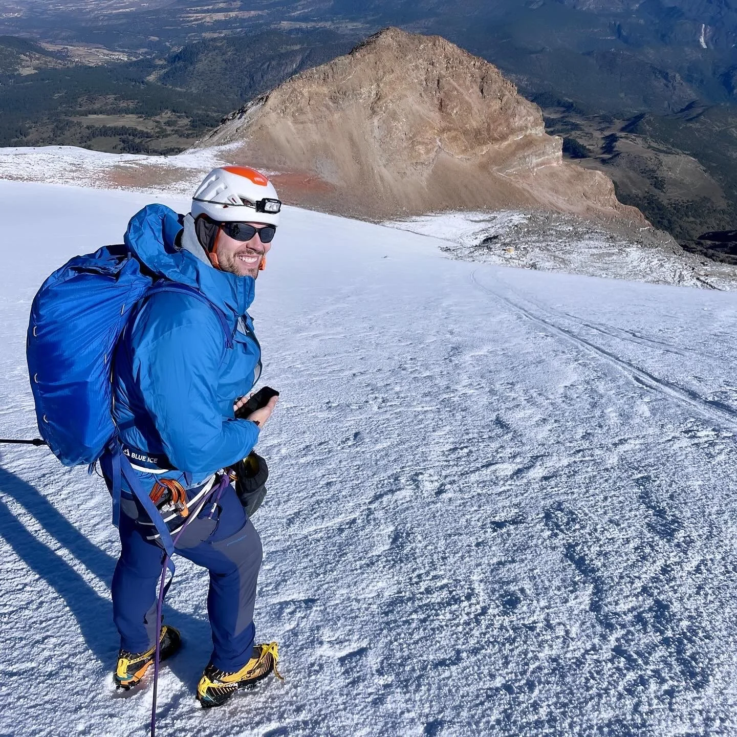 Man in blue winter jacket and pants with hiking gear, snowshoes, and helmet standing on a snow-covered mountain slope, smiling, with a mountainous landscape in the background.