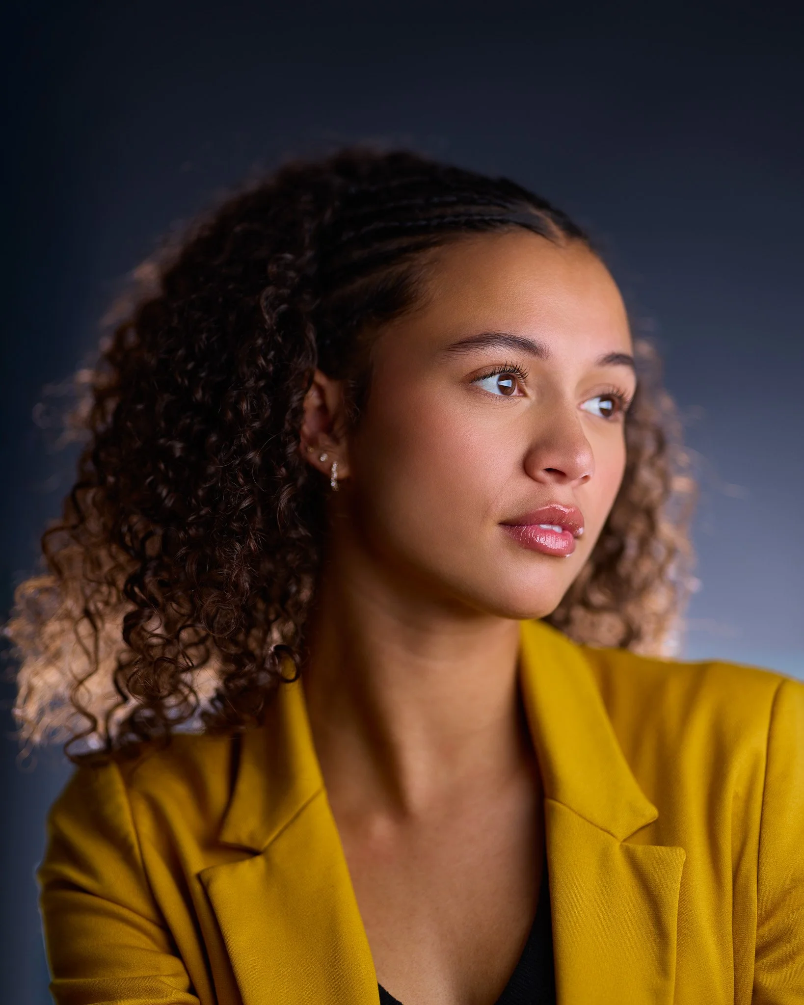 Close-up of a woman with curly brown hair wearing a yellow jacket and earrings, looking to the side against a dark background.