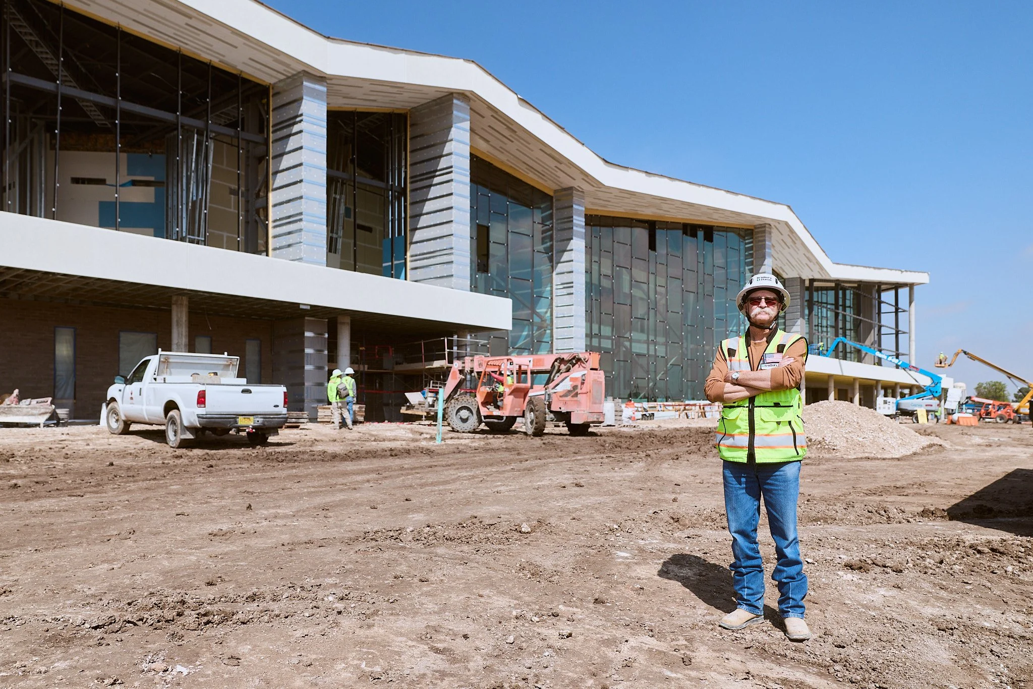 Construction site with a modern building under construction, a worker wearing a hard hat and reflective vest in the foreground, and construction vehicles in the background.
