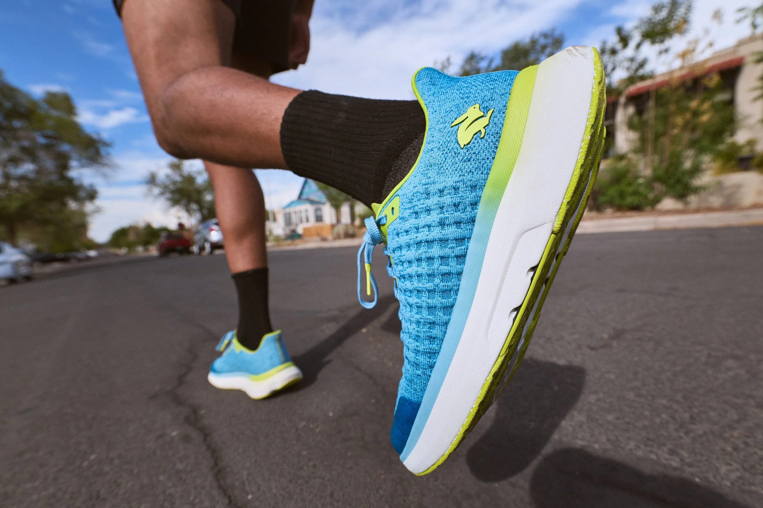 A close-up of a person’s blue running shoe with yellow accents, lifted off the ground during a run outside on a street, with blurred trees, houses, and sky in the background.