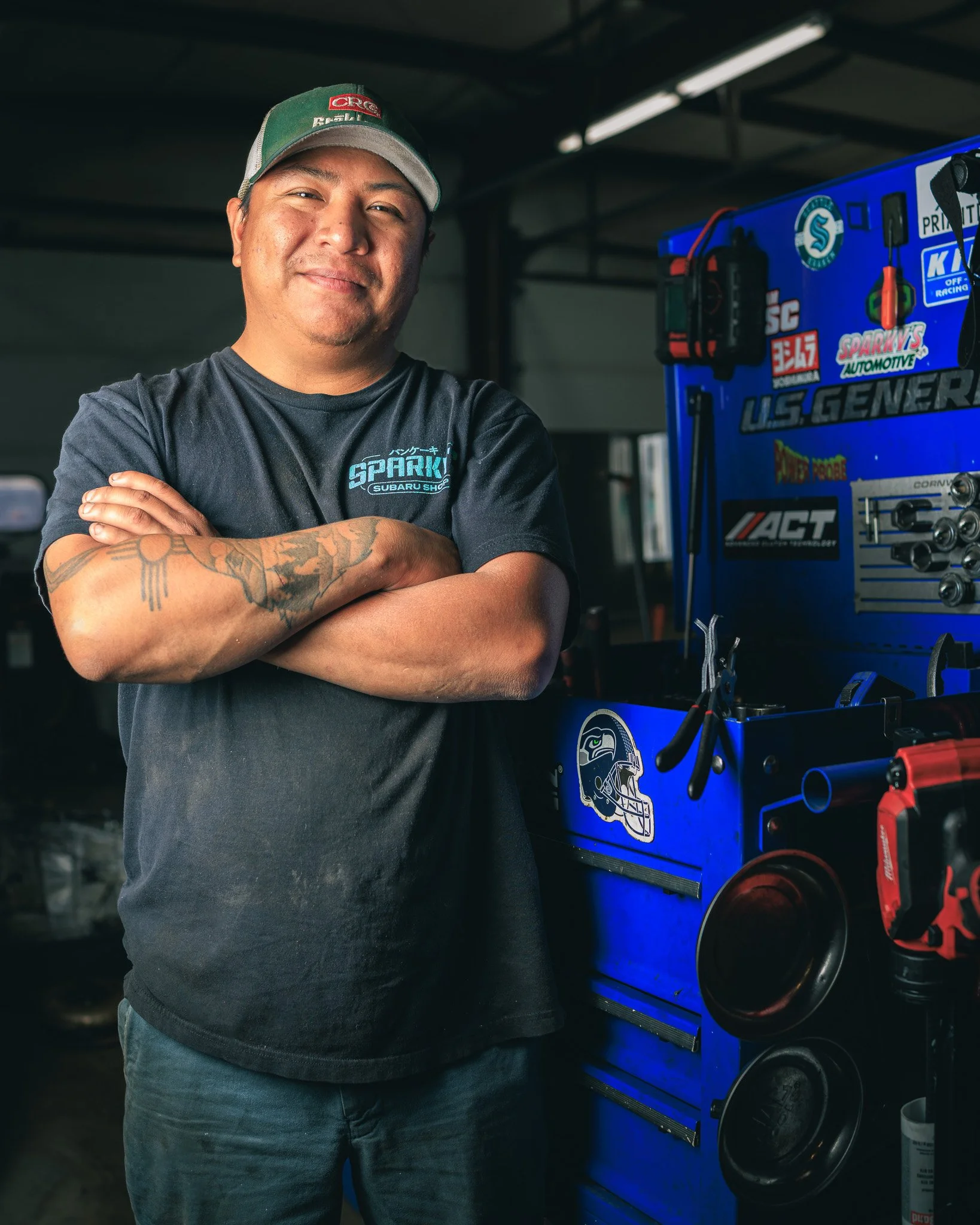 A man with tattoos on his arm stands with his arms crossed, smiling. He wears a dark t-shirt with a Subaru logo and a green cap. Behind him is a blue tool chest with automotive equipment and stickers.
