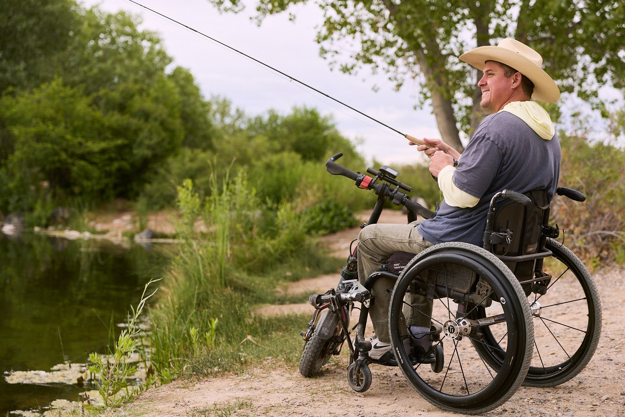 Man in a wheelchair fishing by a pond surrounded by trees.