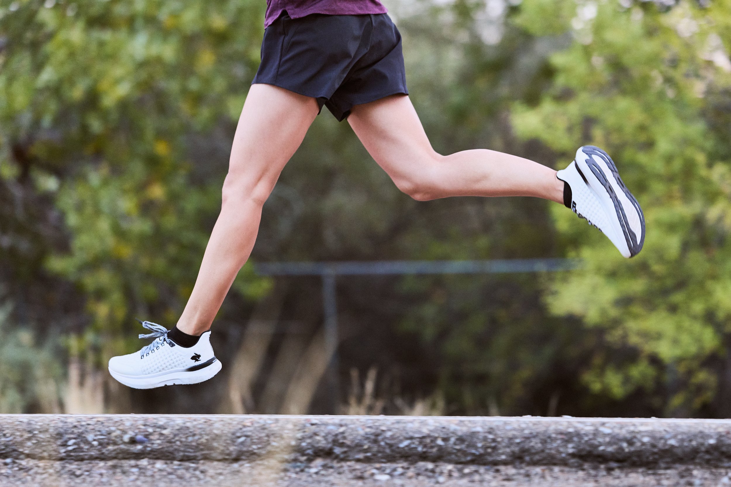 A person running outdoors, wearing black shorts, white running shoes, and a purple top, with trees and greenery in the background.