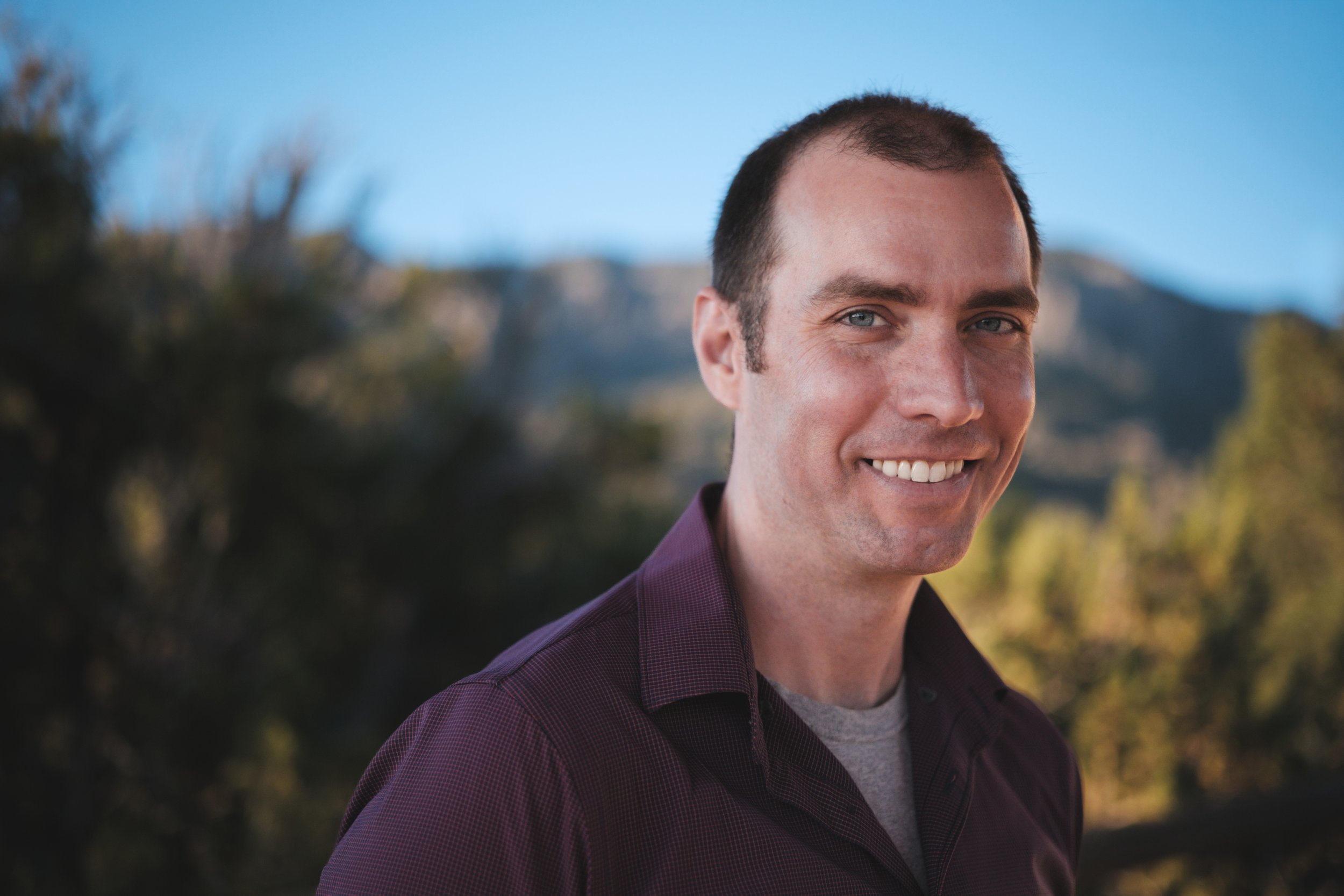 A young man with short dark hair and blue eyes smiling outdoors with a mountain and trees in the background.