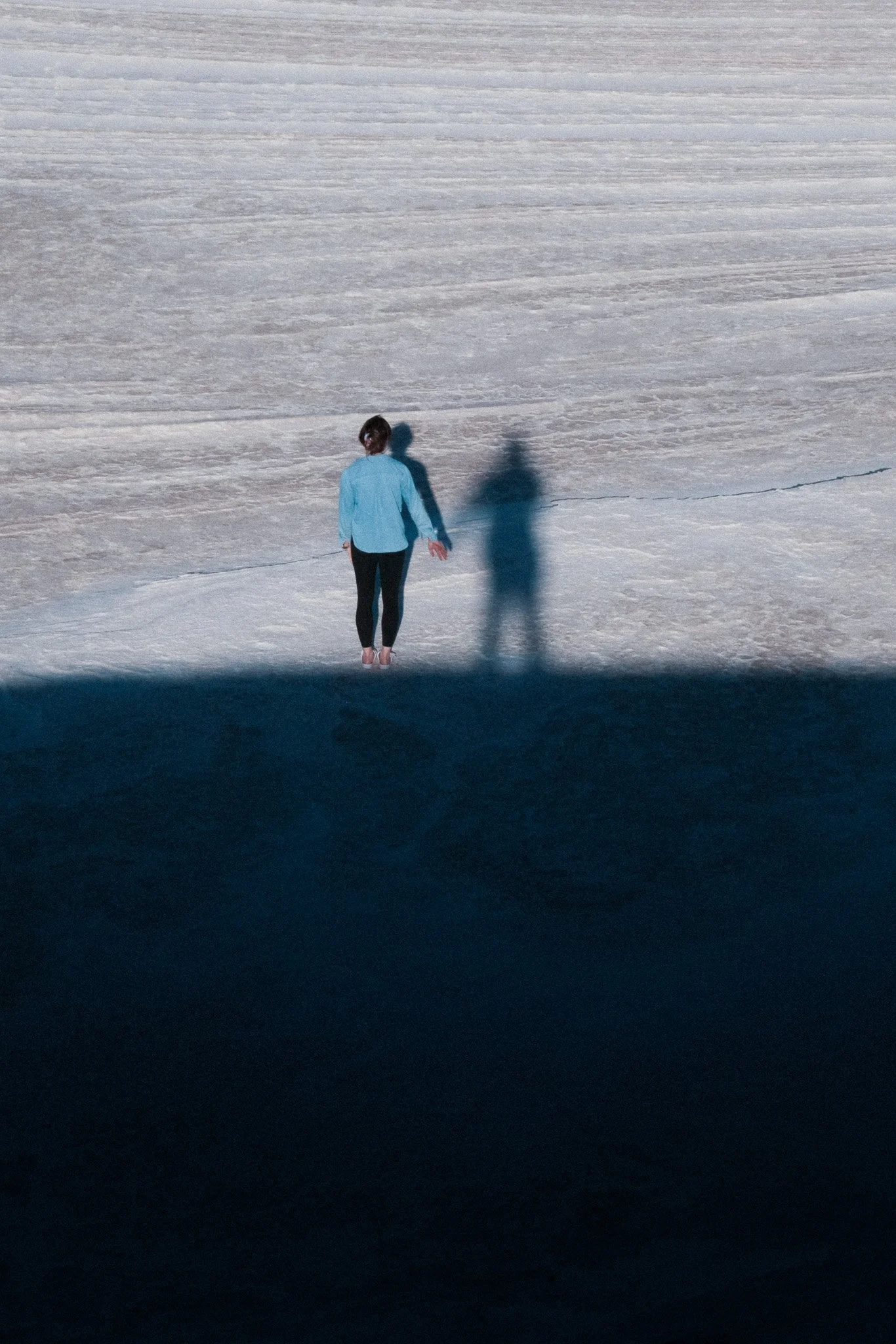 A person in a blue shirt and black pants stands facing away on a snow-covered landscape, casting a shadow.
