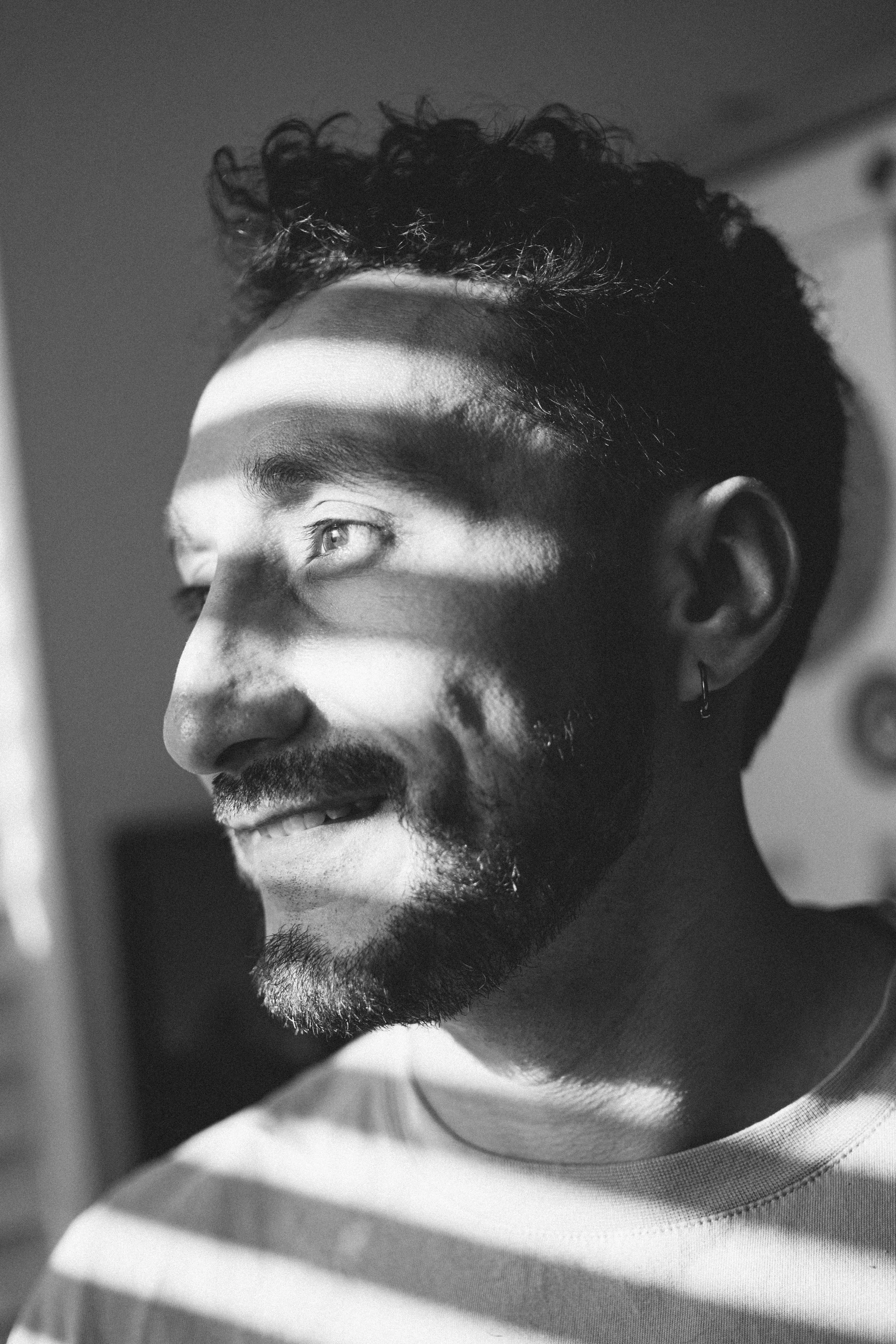 Close-up black-and-white photo of a smiling man with curly hair, a beard, and an earring, looking away, with shadows cast across his face.
