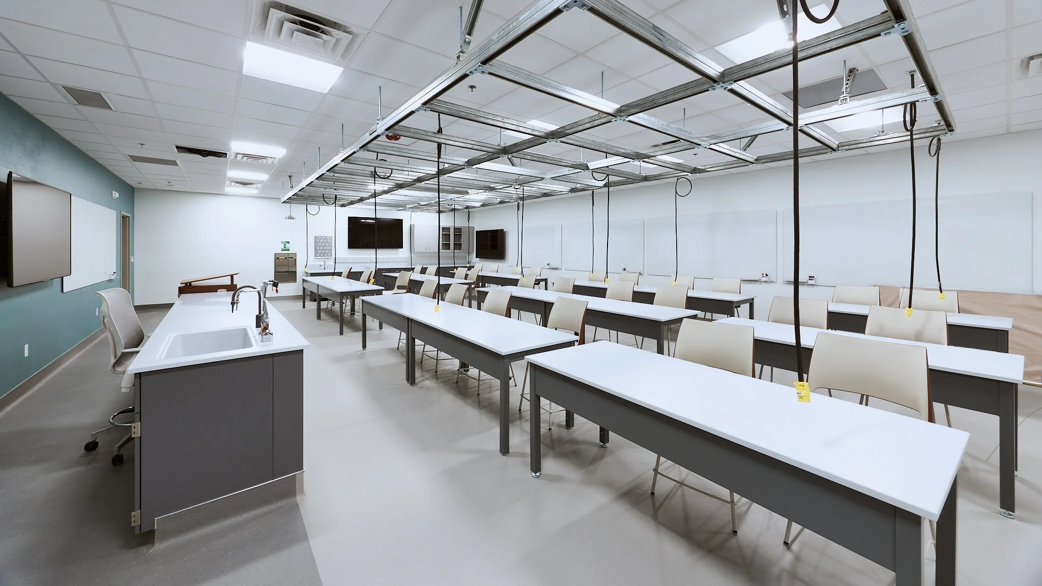 Empty classroom with white desks, beige chairs, whiteboards, multiple monitors, and an overhead metal framework with hanging cables.