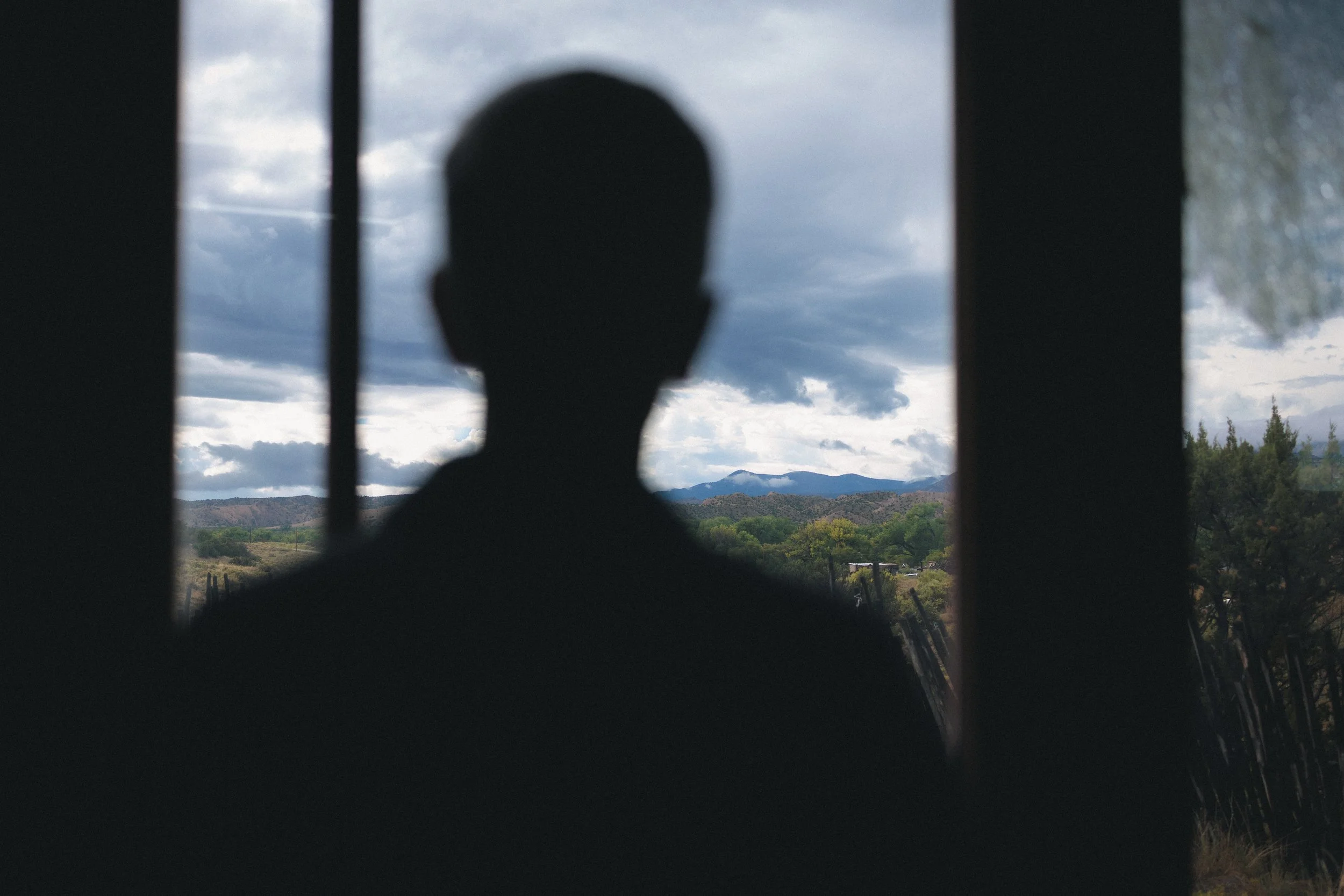Silhouette of a person viewed through a barred window with a landscape of mountains, trees, and cloudy sky in the background.