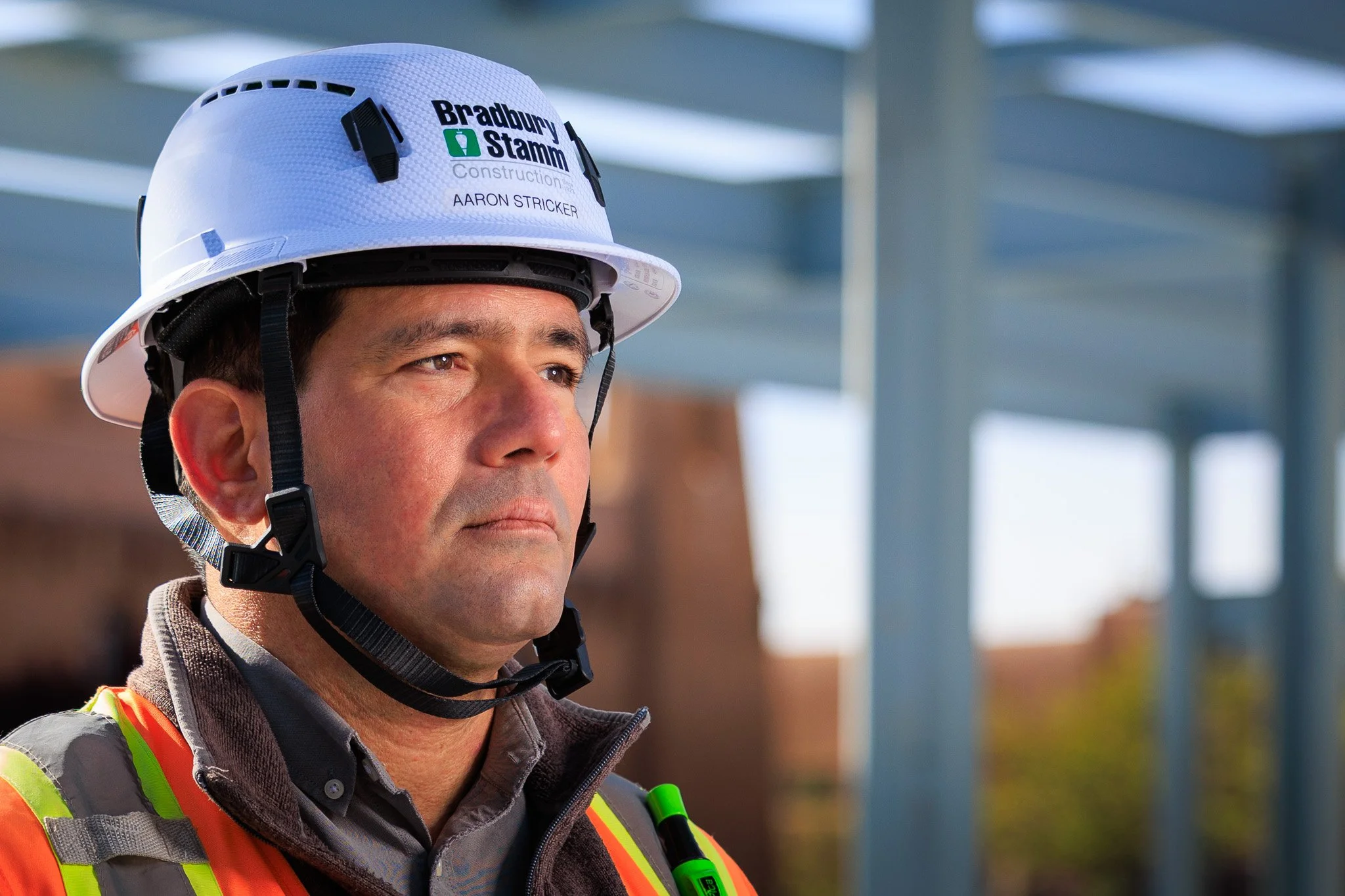 Close-up of a construction worker wearing a white safety helmet with the name tag 'Aaron Stricker' and company logo, wearing a high-visibility safety vest, outdoors on a sunny day.