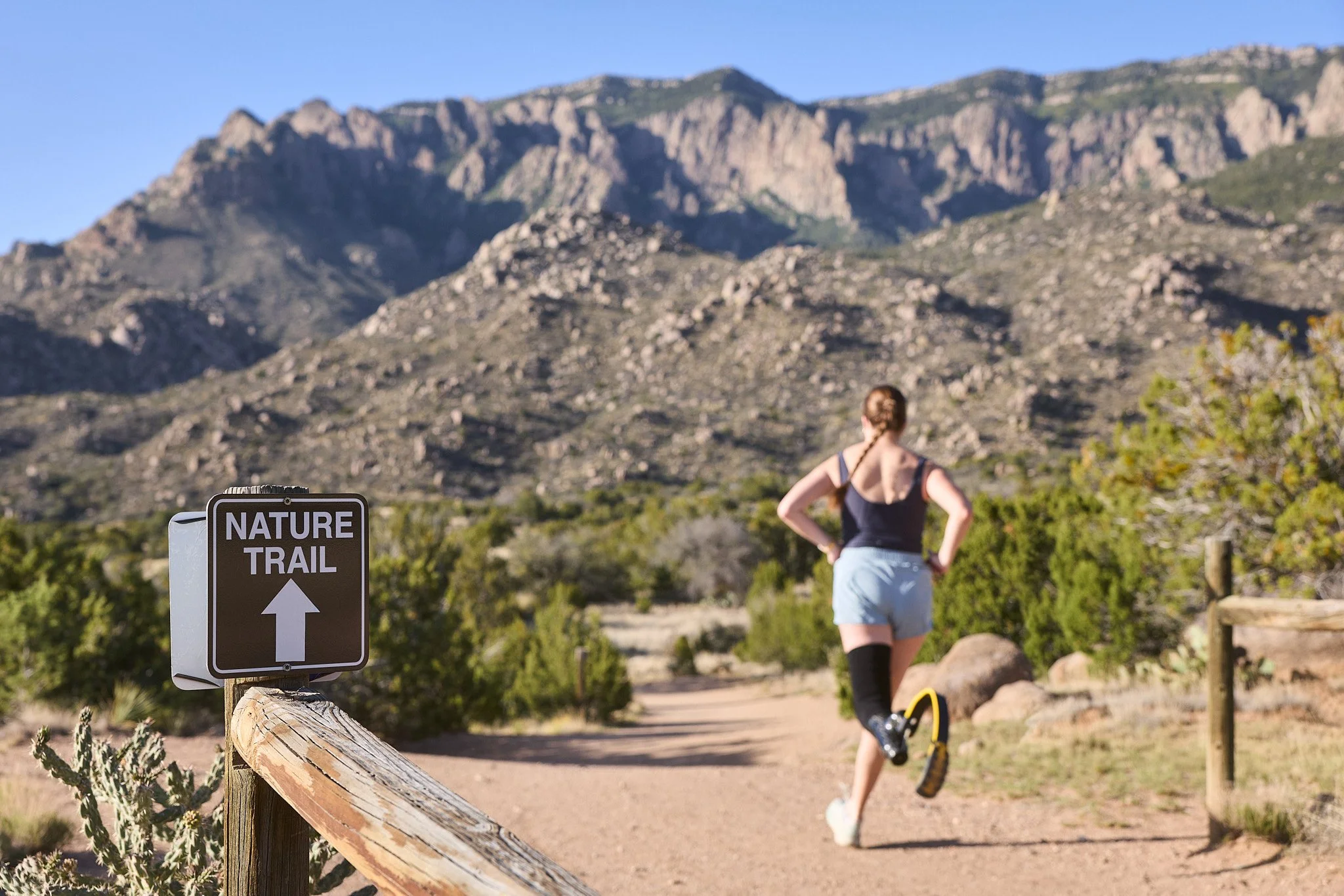 A person with braided hair standing on a nature trail in a mountainous area with rocky peaks in the background. There is a sign that reads 'Nature Trail' with an upward arrow, indicating the trail direction.