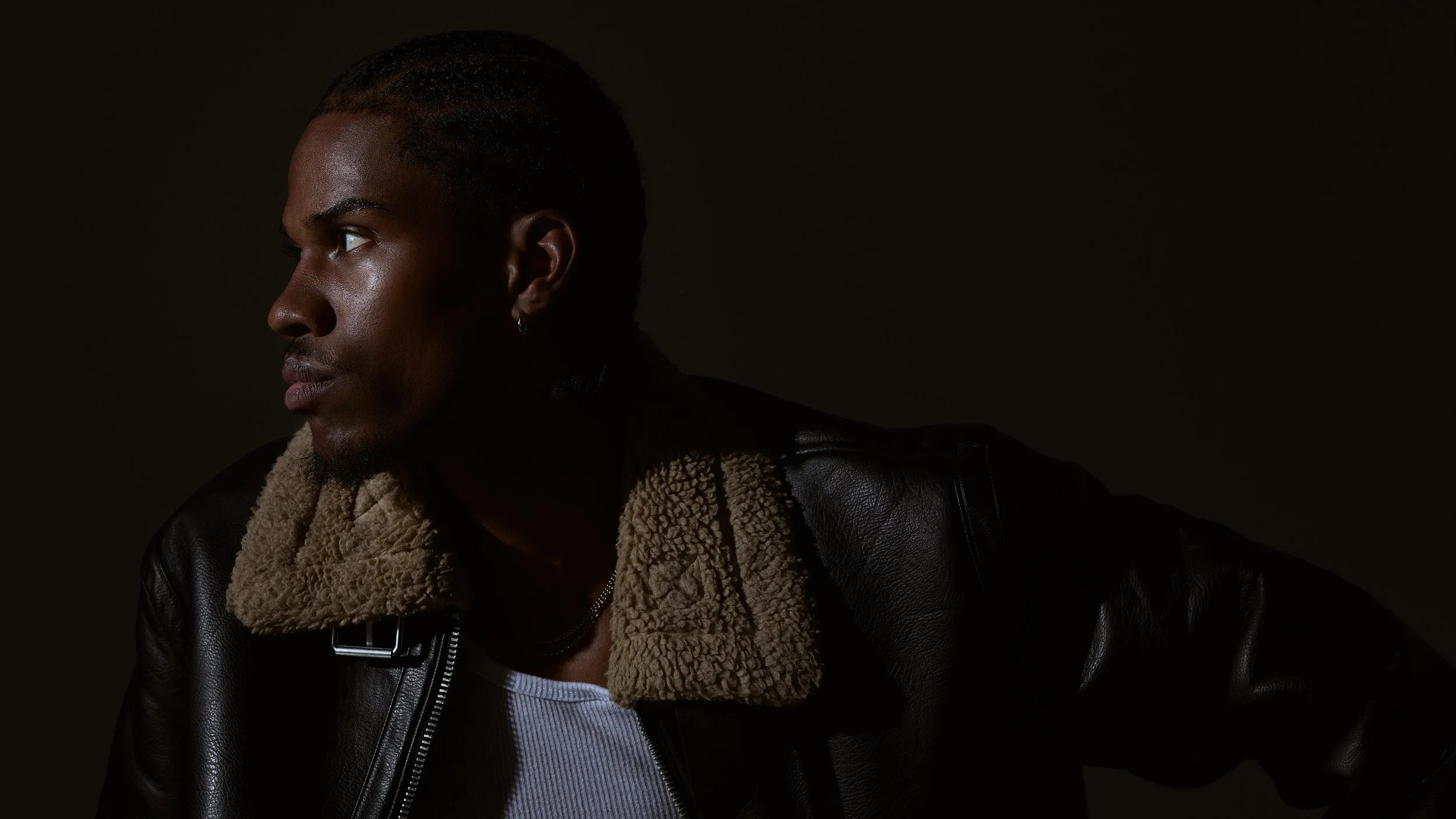 Side profile of a young man with dark skin, short hair, and wearing a black jacket with shearling collar and a white shirt, against a dark background.