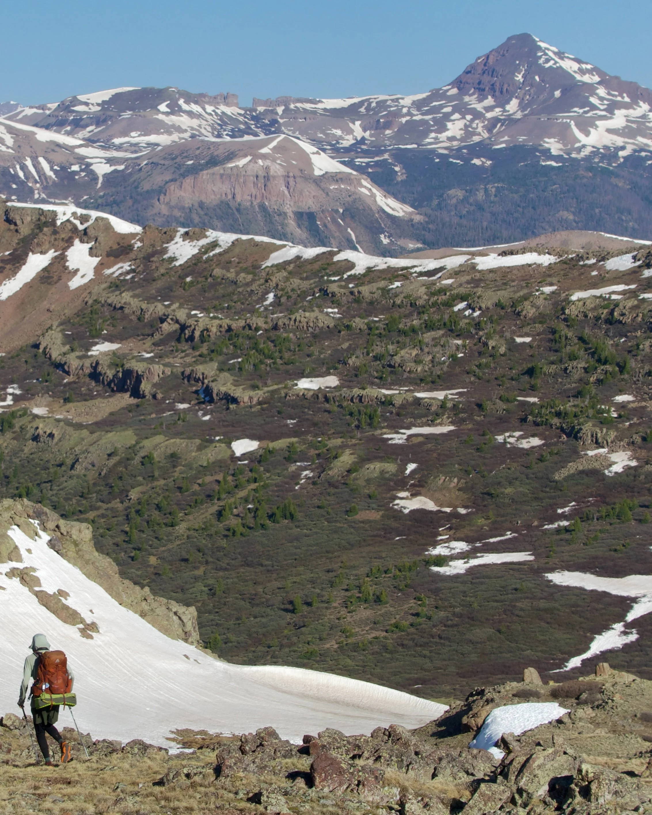 A hiker with a backpack walking on a snowy mountain trail surrounded by rocks and sparse vegetation, with snow-capped mountains and rugged terrain in the background.