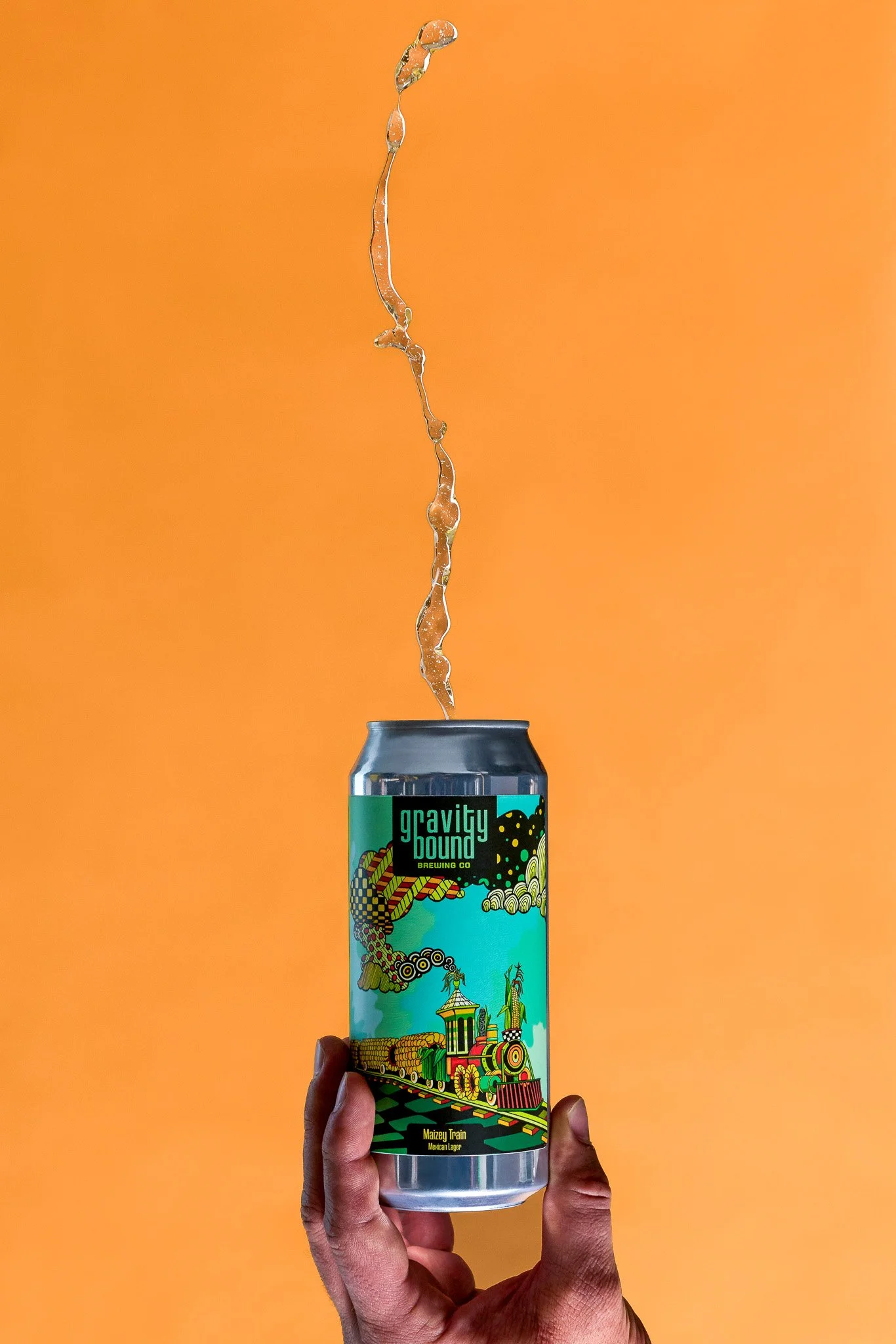 A hand holding a can of Gravity Bound Brewing Co. Maizey Train Mexican Lager with a colorful cartoon-style label featuring a train and cityscape design against an orange background. Liquid is seen overflowing from the top of the can.