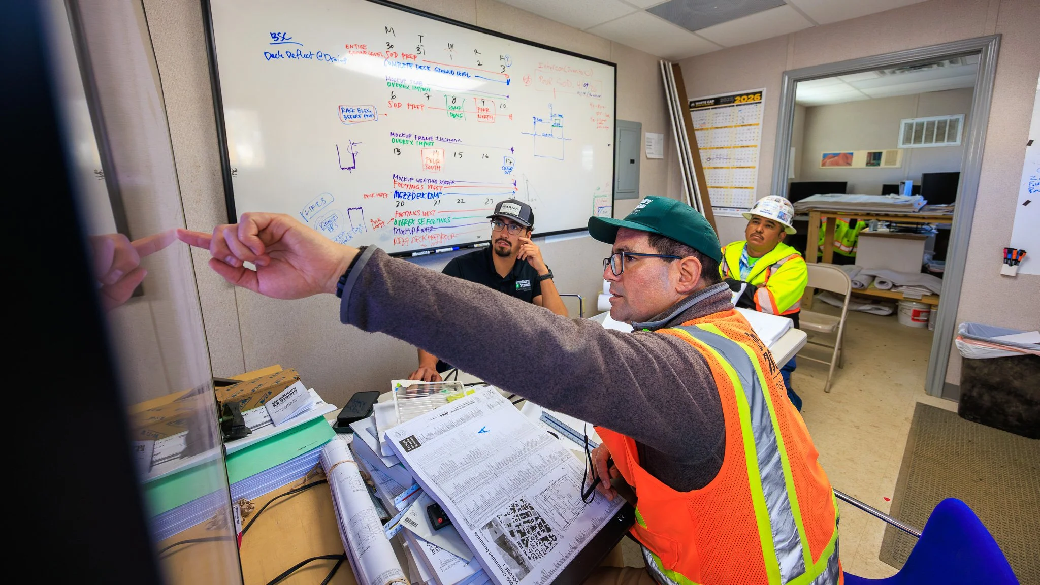 Three men in a construction or engineering office, one in a neon safety vest and hard hat, another in safety gear, and a third man with glasses and a cap, discussing and pointing at a whiteboard filled with notes and diagrams.