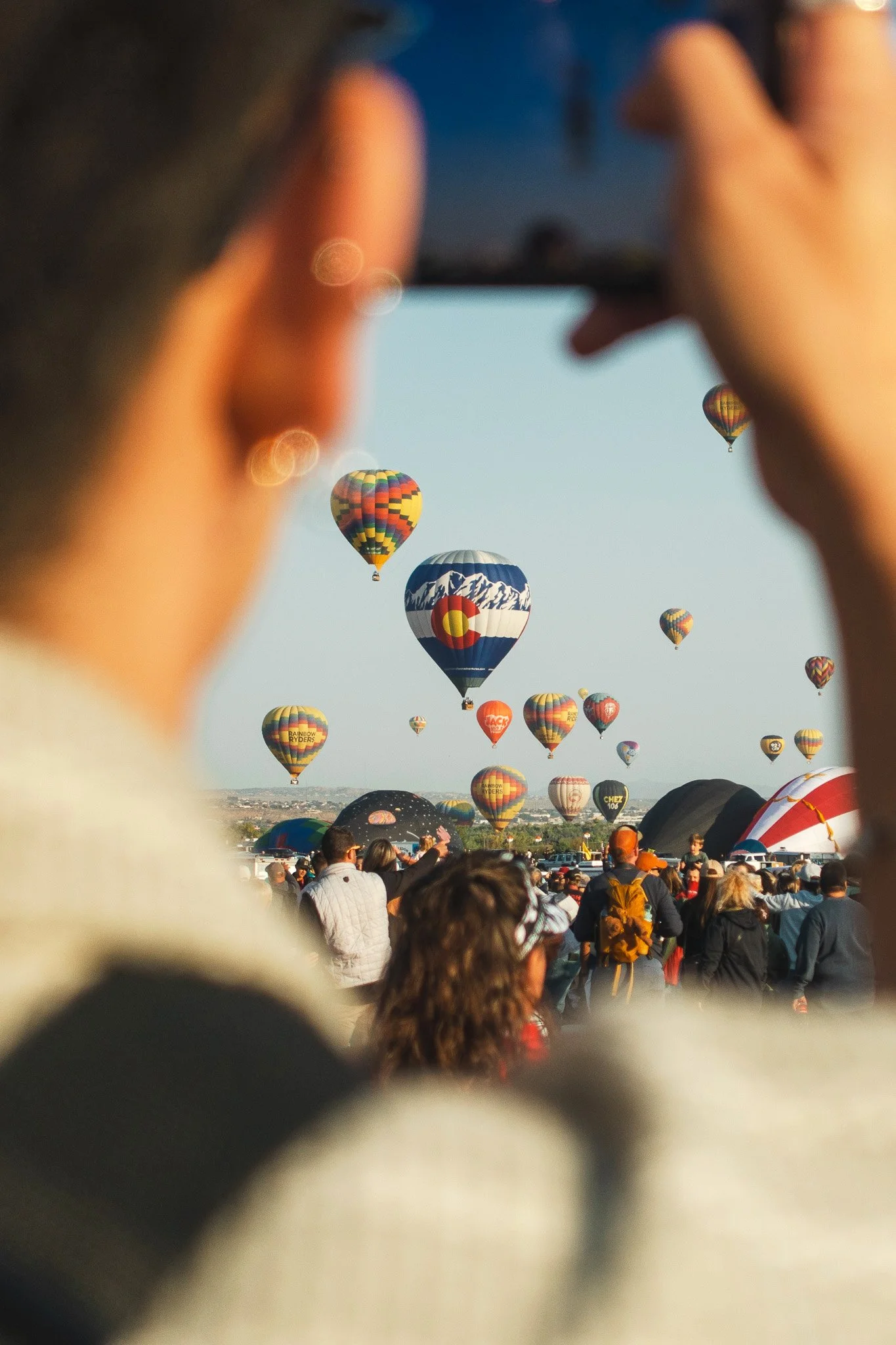 A large crowd of people gathered outdoors with many hot air balloons in the sky, varying in colors and patterns, during a hot air balloon festival.