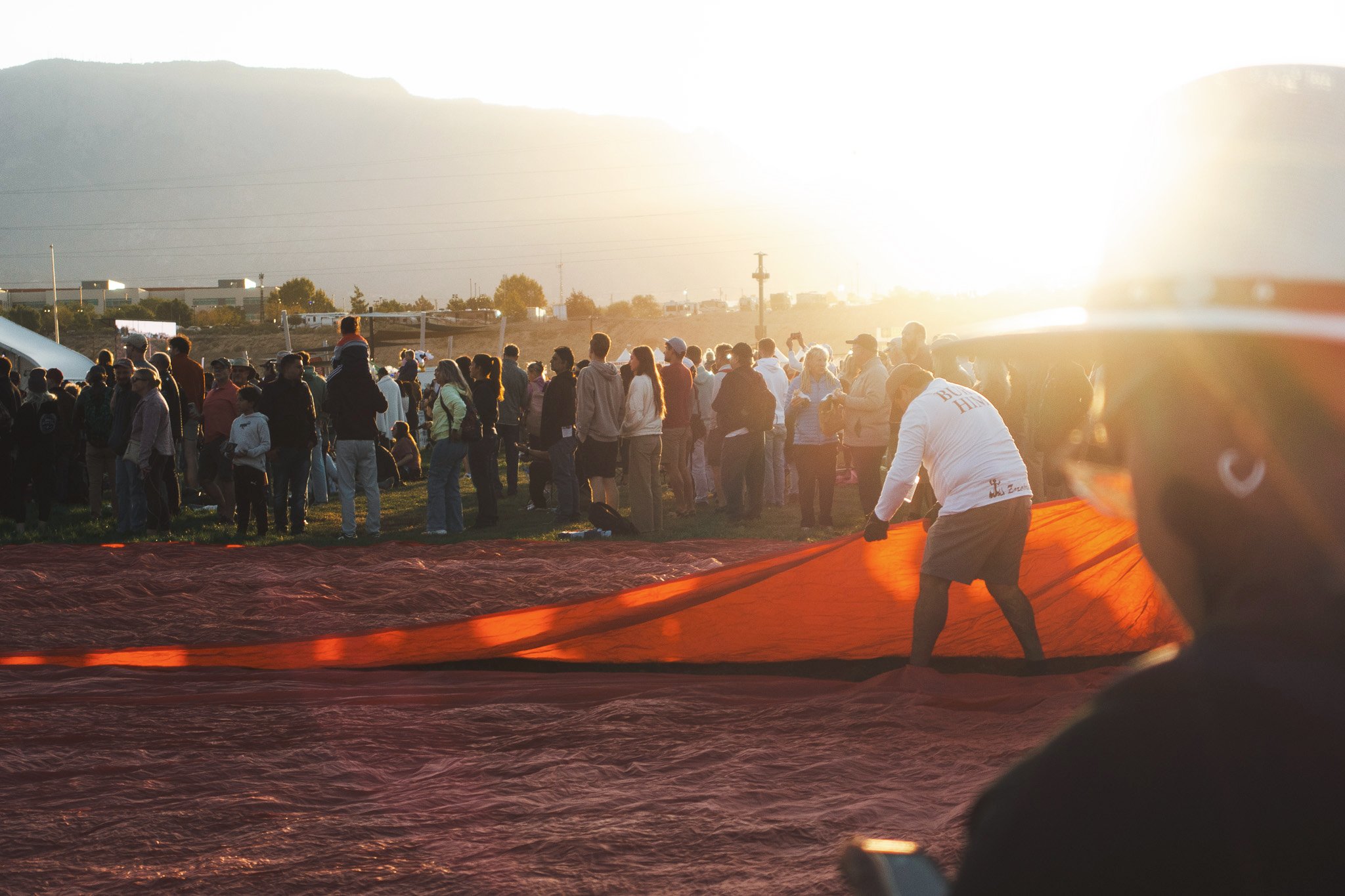A crowd of people gathered outdoors at sunset, some standing and some sitting, with a large red banner or fabric on the ground, and a mountain range in the background.