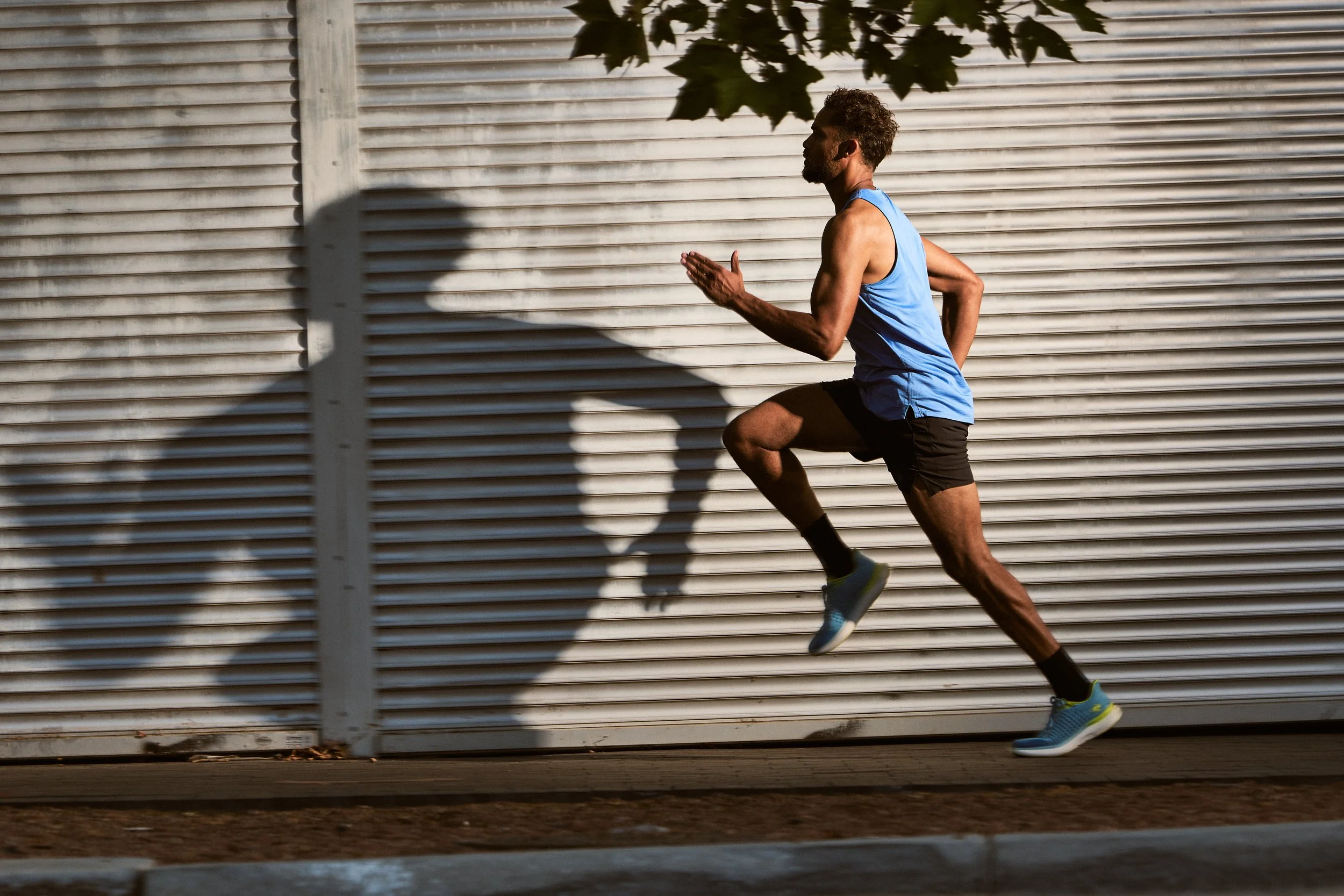 A man running outdoors along a sidewalk, wearing a blue sleeveless shirt, black shorts, and blue running shoes. The background shows a corrugated metal wall and a shadow of a tree, with some leaves overhead.