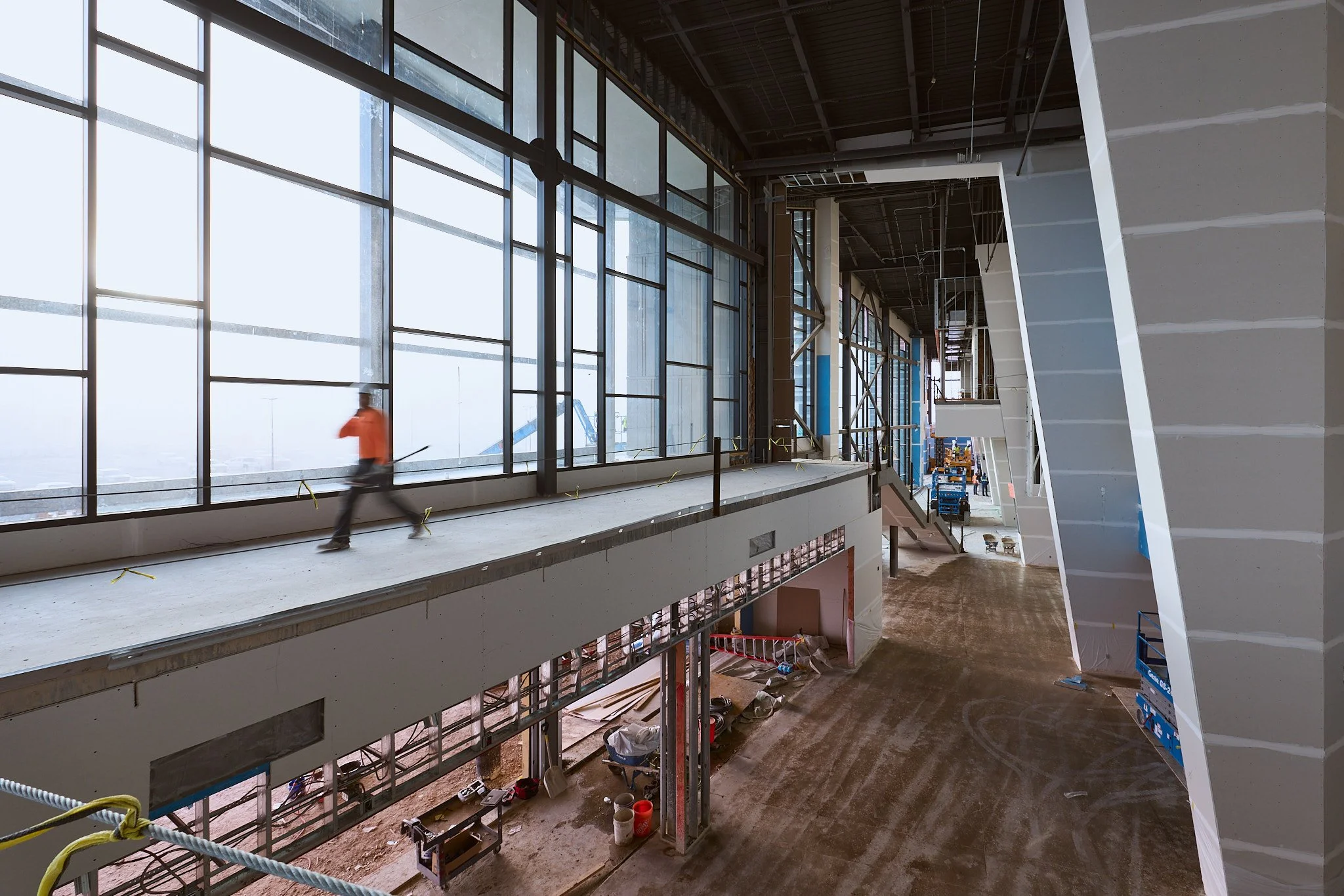 Interior of a building under construction, showing large glass windows, an open area with exposed ceilings, and construction tools and materials on the floor.