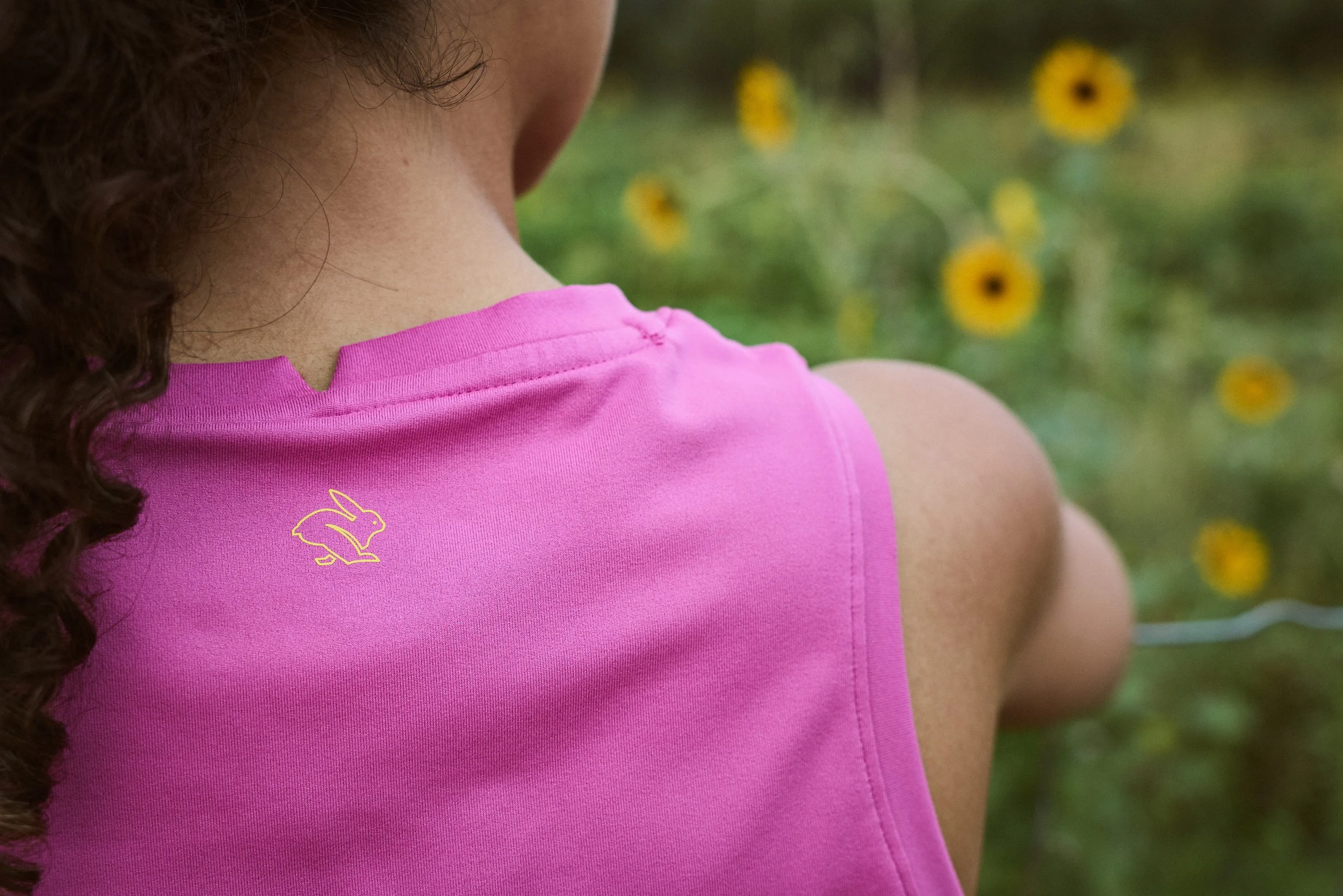 A person with curly hair wearing a pink sleeveless shirt with a small yellow bunny logo on the shoulder, outdoors with yellow flowers in the background.