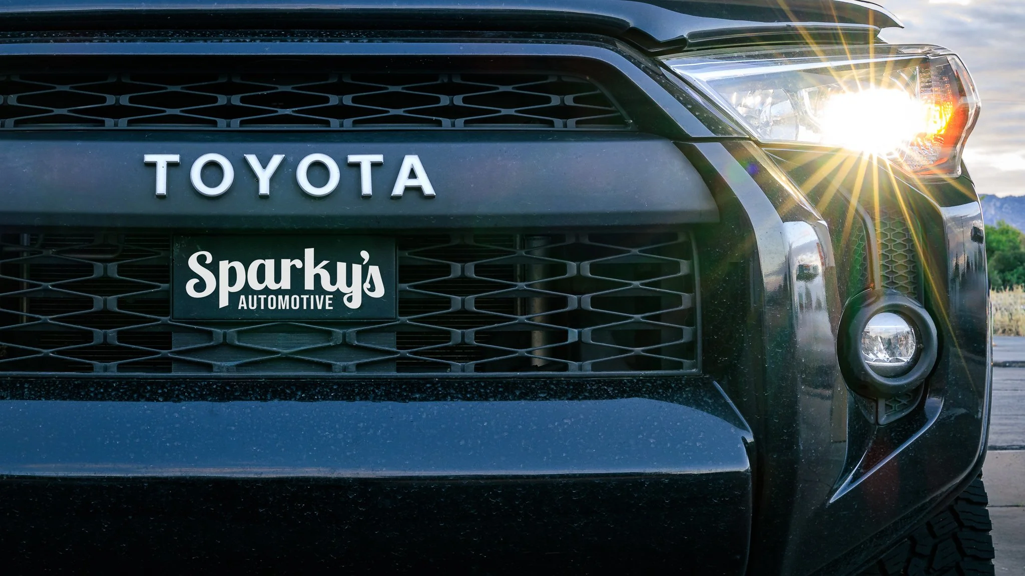 Close-up of the front of a black Toyota vehicle with sunlight reflecting off the headlight, showing the grill and a license plate that reads 'Sky's Automotive'.