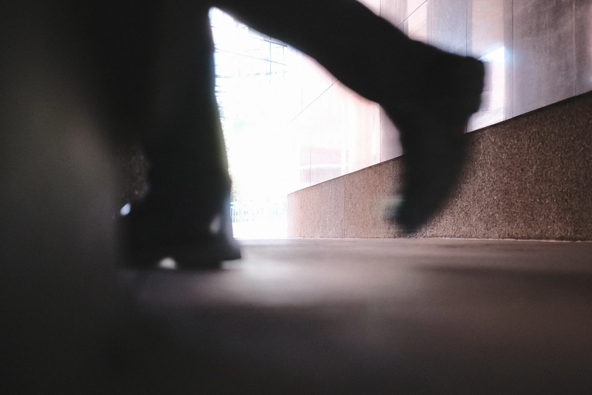 A blurry, out-of-focus photo taken from a low angle shows a modern indoor setting with tiled flooring, large windows, and a person walking away from the camera, partially obstructed by a dark object in the foreground.