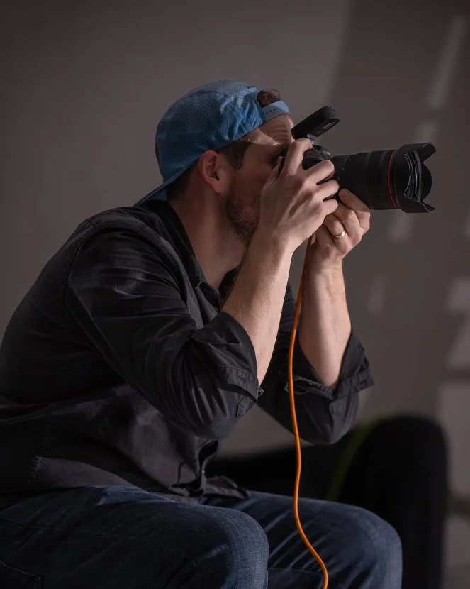 Man wearing a blue cap, sitting and taking a photo with a professional camera, with a grey background.