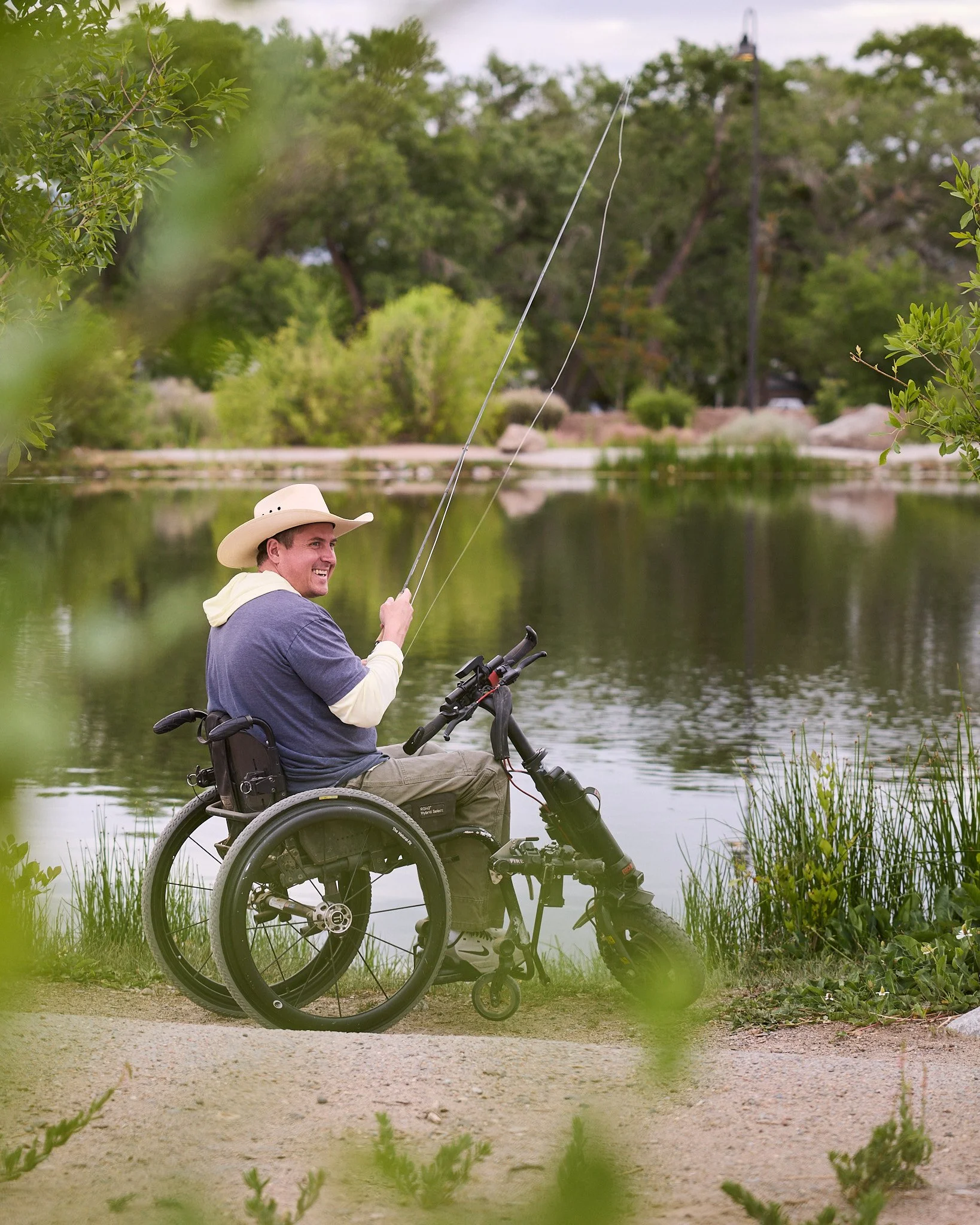 A man in a motorized wheelchair fishing by a lake, wearing a wide-brimmed hat and casual clothing, surrounded by greenery and trees.