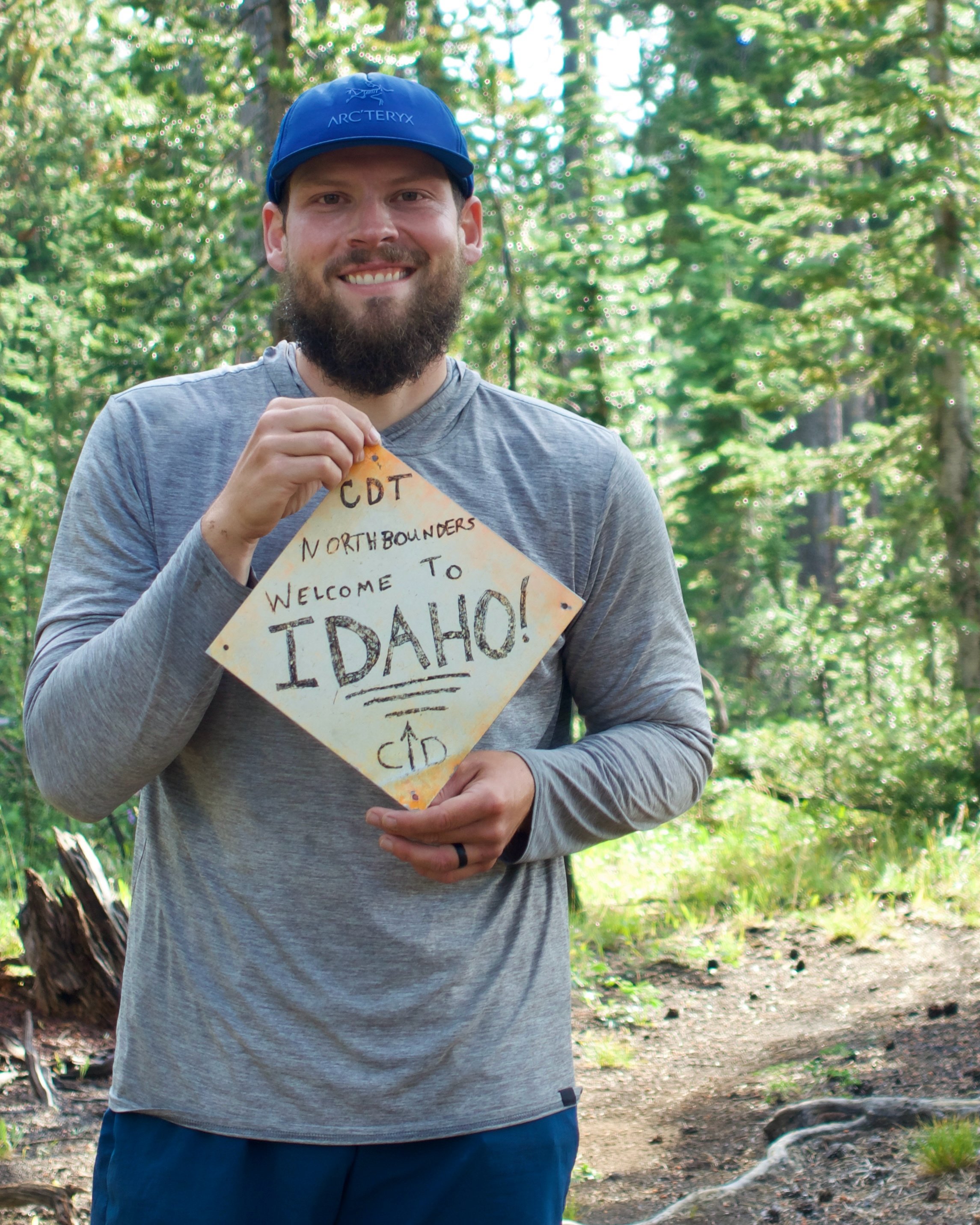 Man with a beard wearing a blue cap and a gray long-sleeve shirt, holding a handwritten sign that reads 'CDT Northbounders Welcome to Idaho!' in a forest setting.