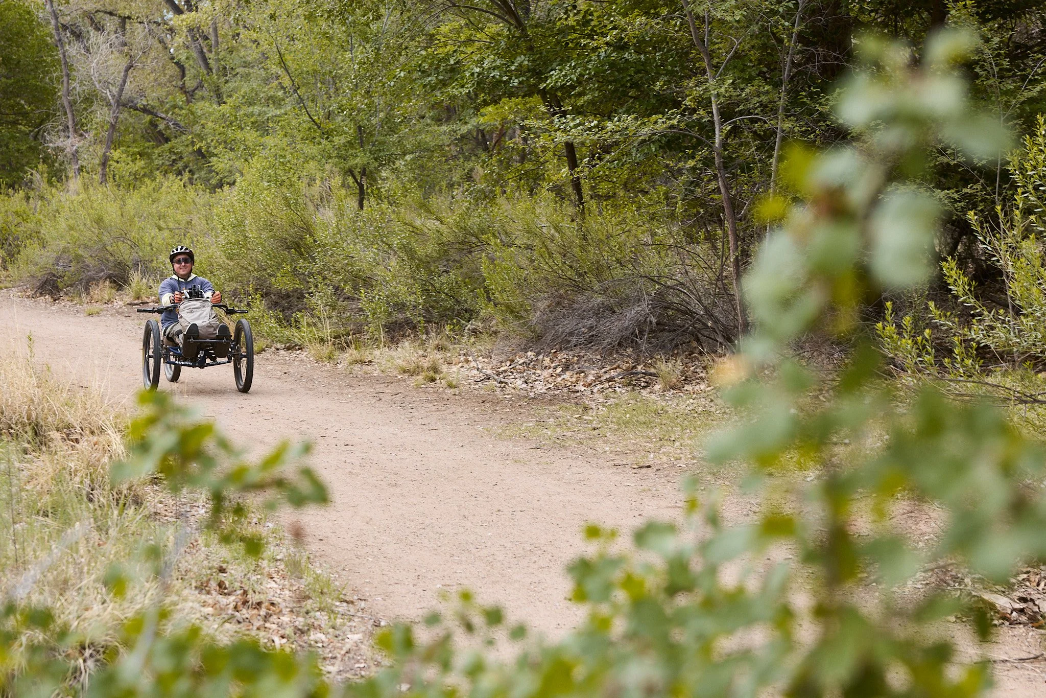 A person riding a handcycle on a dirt trail surrounded by green trees and bushes.